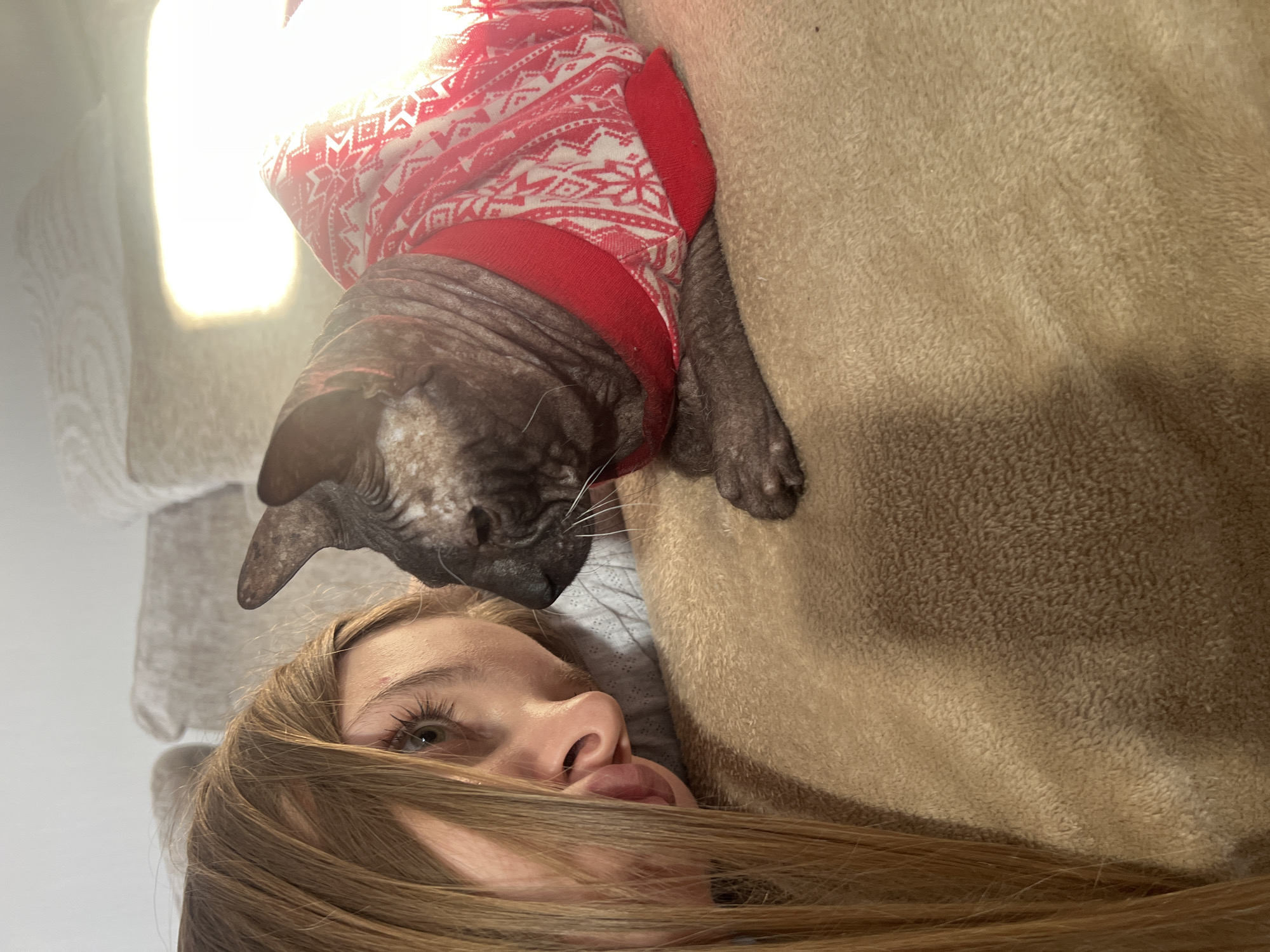 A woman is lying down, with a hairless cat wearing a red and white sweater lying next to her. The background appears to be a tan blanket. The photo was not taken in Votkinsk, Russia.