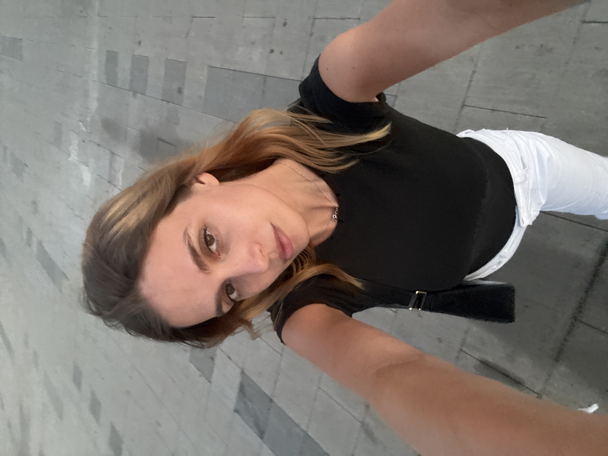 This is a high-angle, selfie-style photograph of a young woman with blonde hair and brown eyes. She is wearing a black t-shirt and white pants, and a black bag is slung over her shoulder. The woman is looking directly at the camera with a neutral expression. She is standing on a paved outdoor area with a pattern of rectangular tiles. The background suggests an urban environment, consistent with a location in Belgrade, Serbia, potentially an outdoor public space like a square or promenade.