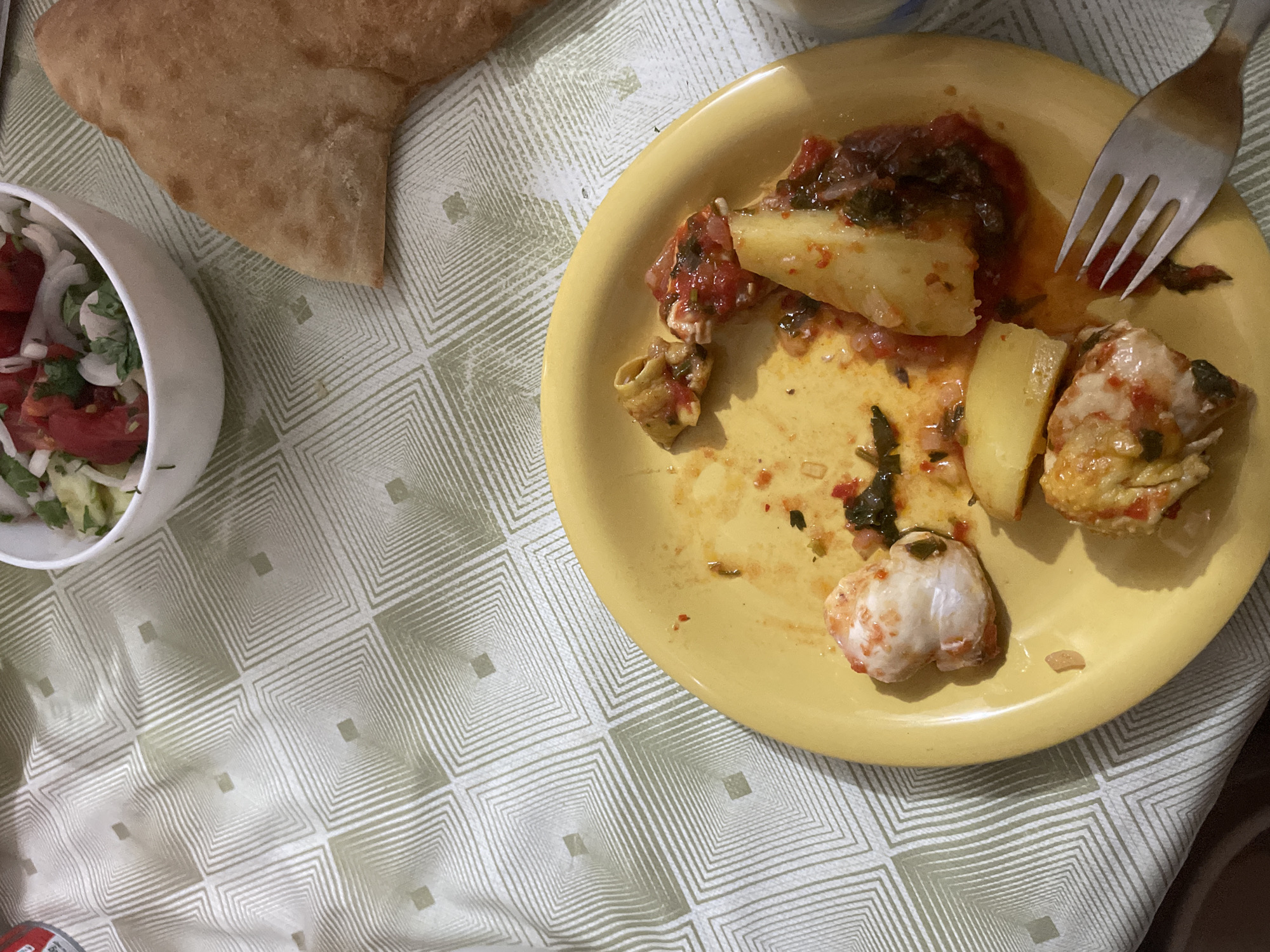 The image shows a close-up of a yellow plate with leftover food, a white bowl of salad, and a piece of flatbread on a tablecloth with a geometric pattern. The plate contains what appears to be a seafood dish with chunks of white meat, potato wedges, and a red sauce with herbs. A silver fork rests on the right side of the plate. In the background, a white bowl holds a salad made with chopped tomatoes, onions, and greens. A piece of golden-brown flatbread is positioned to the right of the bowl. The setting appears to be a casual meal, possibly at a restaurant or home in the city of Tsalmiani, Georgia, given the context. The lighting suggests an indoor setting, perhaps during the day or with artificial light.