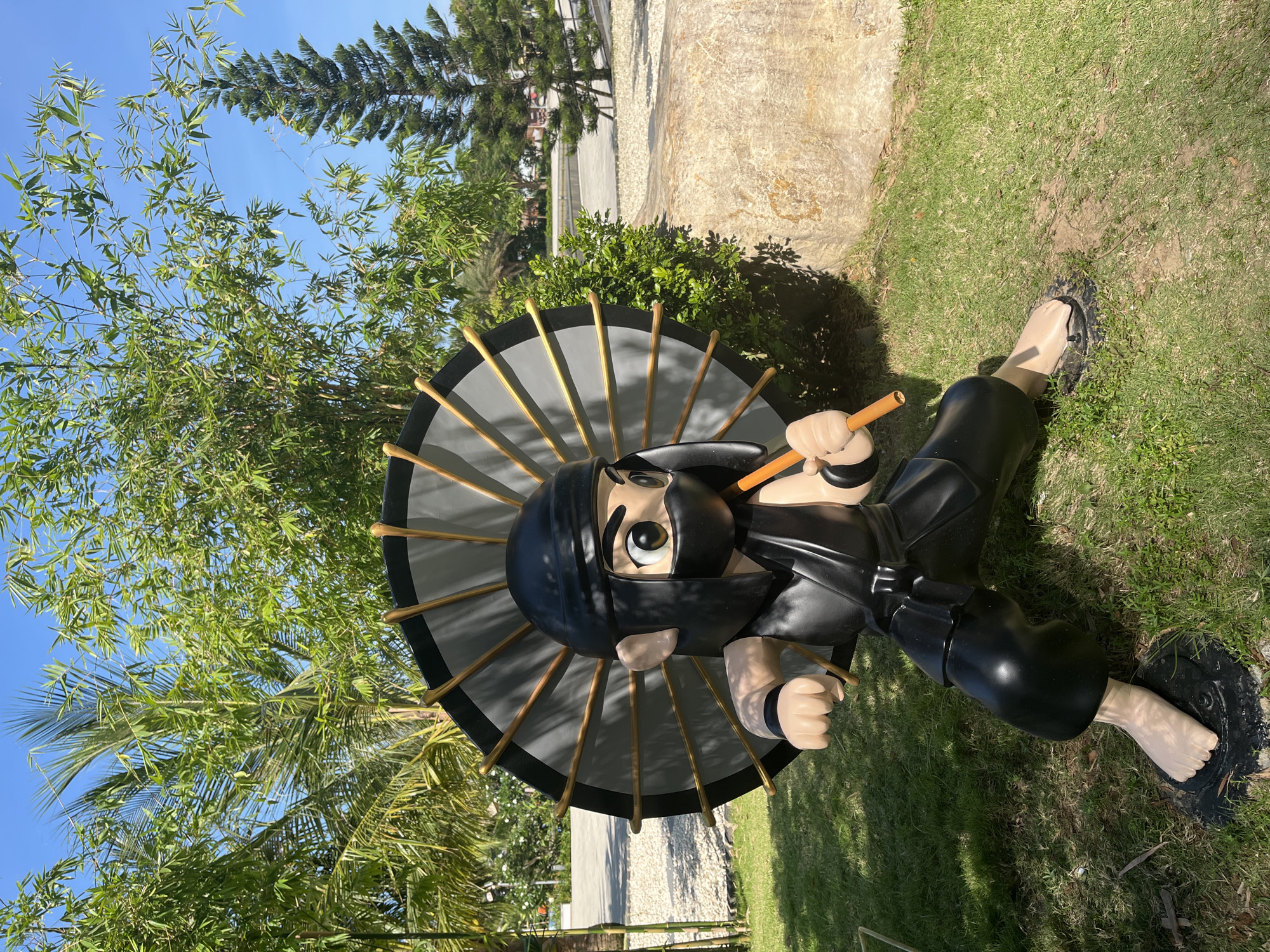An outdoor photograph, taken on a clear and sunny day, features a large, cartoonish statue of a ninja. The ninja statue is predominantly black, depicting a figure in a traditional ninja outfit with a black head covering and a mask obscuring the lower half of its face. One large, round, light-colored eye is visible, giving the statue a whimsical or curious expression.
The statue is posed in a relaxed, sitting or reclining position on a patch of green grass. Its hands and one exposed foot are a light, pale skin tone. The ninja holds a large, open umbrella above its head with its left hand. The umbrella is grey with a black rim and prominent golden-colored spokes. In its right hand, the ninja clutches a wooden stick or staff.
The setting is outdoors, characterized by lush green foliage. Directly behind and surrounding the statue are various trees, including tall bamboo stalks with slender leaves and parts of a palm tree visible on the left. In the background, under a bright blue sky, more trees, possibly pine-like, and light-colored concrete or stone ground are visible. Distinct shadows cast by the trees and the statue indicate bright sunlight, suggesting it is daytime, likely late morning or early afternoon. The presence of bamboo and palm trees, combined with the location context of Đà Nẵng, Vietnam, points to a tropical or subtropical environment. There is no visible text in the image, and the statue appears to be a permanent outdoor decorative installation.