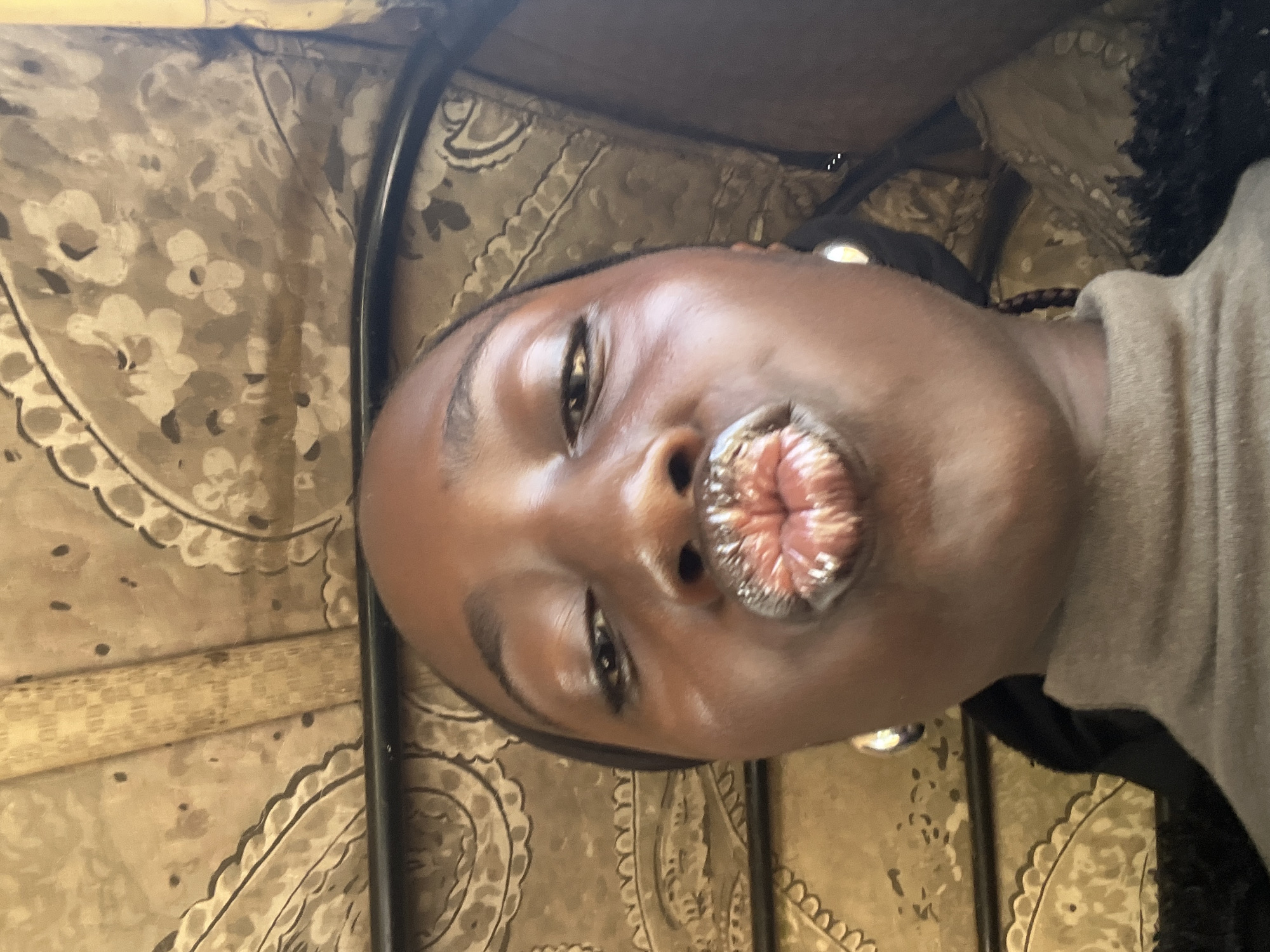 In a selfie taken inside a vehicle in Galadimawa, Nigeria, a young Black woman with dark skin and dark eyes poses with her lips puckered for a kiss. She is wearing a light brown turtleneck sweater and silver hoop earrings. The background appears to be the interior of a vehicle, with patterned fabric upholstery and a metal bar. The lighting is natural and casts a soft glow on her face.