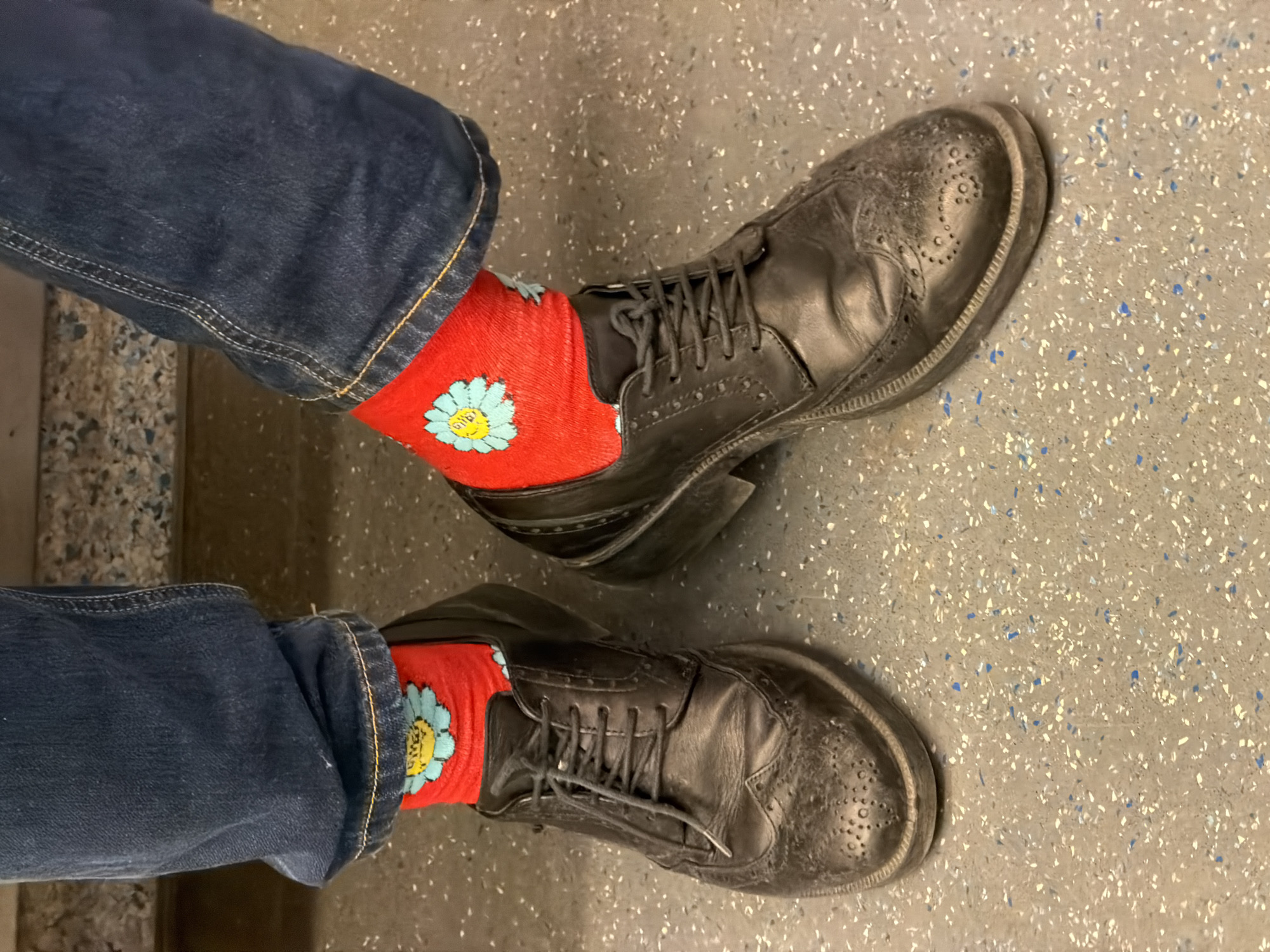 The image shows a pair of legs wearing denim jeans, red socks with a blue flower design, and black leather lace-up shoes. The legs are positioned on a speckled grey floor. The setting is not definitively identifiable as Moscow, Russia, and the information should not be assumed to be true.