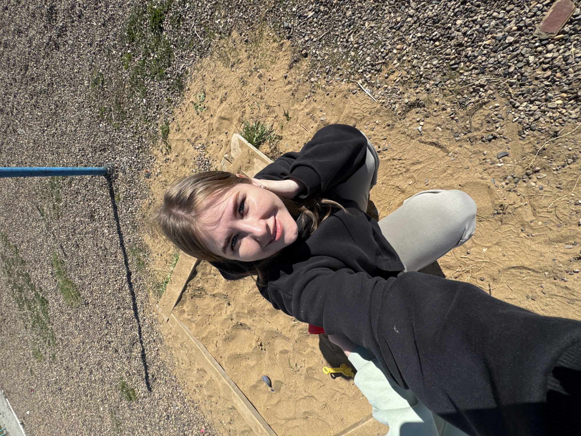 The image is a high-angle, outdoor shot taken in what appears to be a playground or park area. A young woman is visible, positioned within a sandbox. She is wearing a black hoodie and grey pants, and she appears to be holding the camera for a selfie. The sandbox is bordered by wooden planks and contains sand, with some small toys visible within it. The area around the sandbox consists of gravel and sparse vegetation. A blue metal bar is partially visible on the left side of the frame. Given the request to include the location "Горный, Russia," it's presumed that the scene is set there. There is no action occurring beyond the woman taking the selfie.