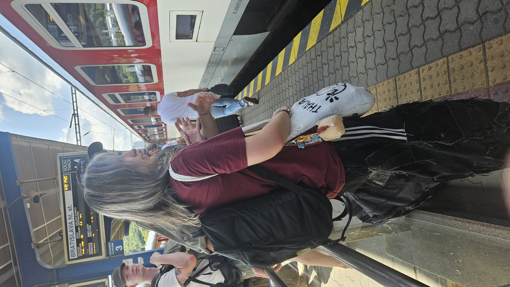 In Bratislava, Slovakia, at a train station platform, a person with gray hair is standing and speaking. They are wearing a maroon t-shirt and black pants. The person has a black backpack on and is holding a white tote bag with a keychain attached. Other people are visible in the background, including a person in a white shirt walking away from the camera, and another person with blond hair and a black cap looking towards the left. A red and white train is parked on the tracks next to the platform. A digital sign above the platform displays "BRATISLAVA hl. st." and "KOLAJ TRACK 4", indicating the location and track number. The platform is paved with interlocking bricks and has yellow and black tactile paving along the edge. The sky is blue with some clouds.