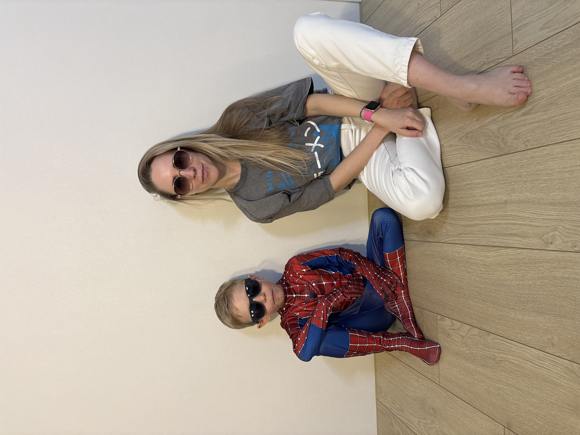 A woman and a child in a Spider-Man costume are sitting on a light-colored wooden floor in front of a white wall. The woman is wearing sunglasses, a grey T-shirt, and white pants. The child is wearing sunglasses and a full Spider-Man suit. The location is implied to be Chaikovsky, Russia.
