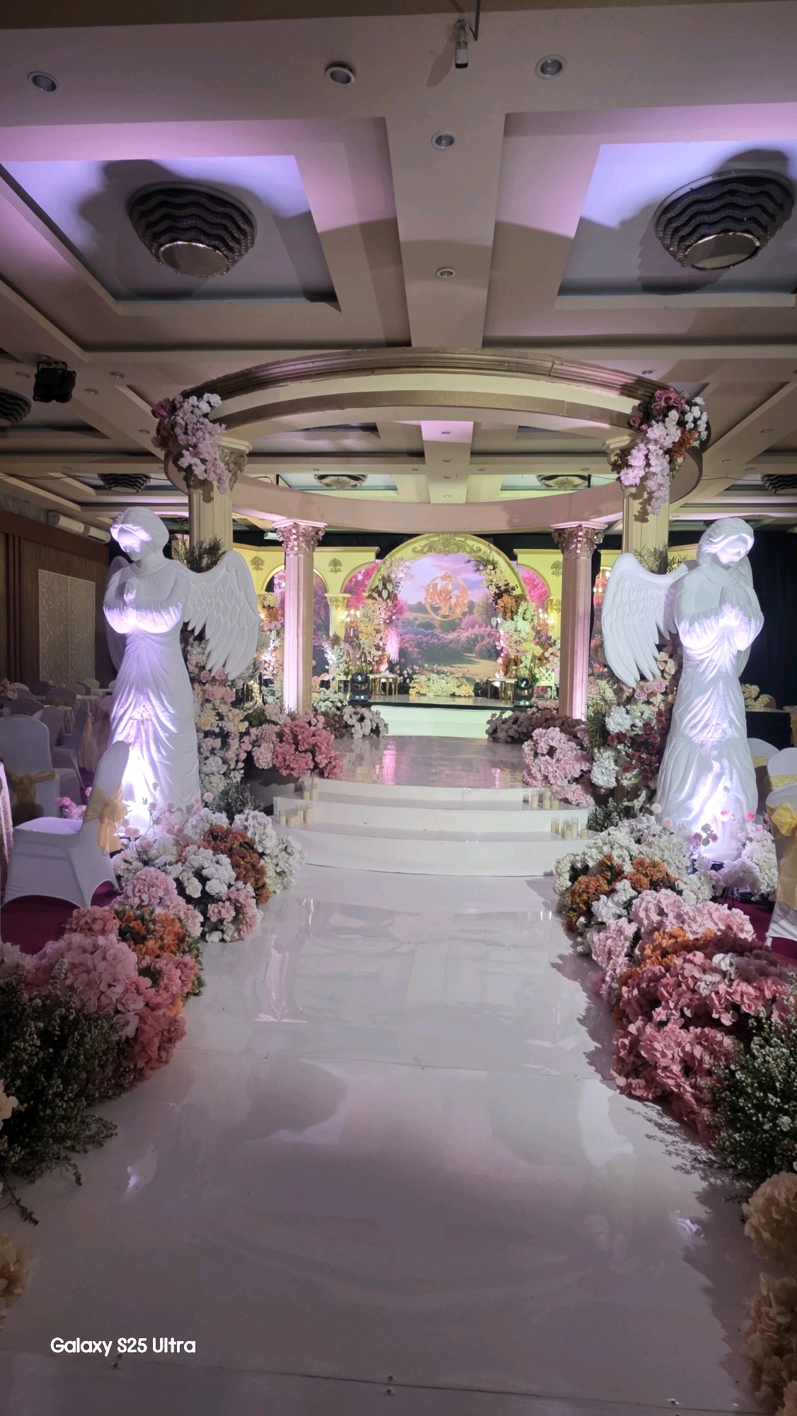 The image depicts a lavish wedding or event venue decorated for a ceremony. The scene is set indoors, likely in a banquet hall, with a grand archway at the far end serving as a backdrop. This backdrop features a painted mural with a pastoral or mythological scene, adorned with abundant floral arrangements in shades of pink, white, and peach. Flanking the entrance to the backdrop area are tall, white columns topped with floral displays.
In the foreground, a white, reflective aisle leads towards the backdrop. On either side of the aisle, large, white statue-like figures of angels stand prominently. These angel figures are draped and have wings, with their hands clasped or in a prayerful pose. They are also surrounded by generous arrangements of flowers matching those at the backdrop.
The aisle itself is wide and appears to be made of a reflective material, possibly polished white flooring or a temporary covering. Steps lead up to a raised platform in front of the archway. The ceiling features modern recessed lighting and decorative elements that cast a pinkish-purple hue over the scene, enhancing the romantic ambiance.
Chairs, likely for guests, are visible on the sides of the hall, some draped with white covers and adorned with yellow sashes. The overall impression is one of opulence and romantic elegance, typical of a high-end wedding setup. The "Galaxy S25 Ultra" watermark in the bottom left corner indicates the device used to capture the image.
While the image itself does not provide direct visual cues for the specific location within Kecamatan Pati, Indonesia, this type of elaborate indoor event styling is commonly found in established event venues or hotels in urban and semi-urban areas.
