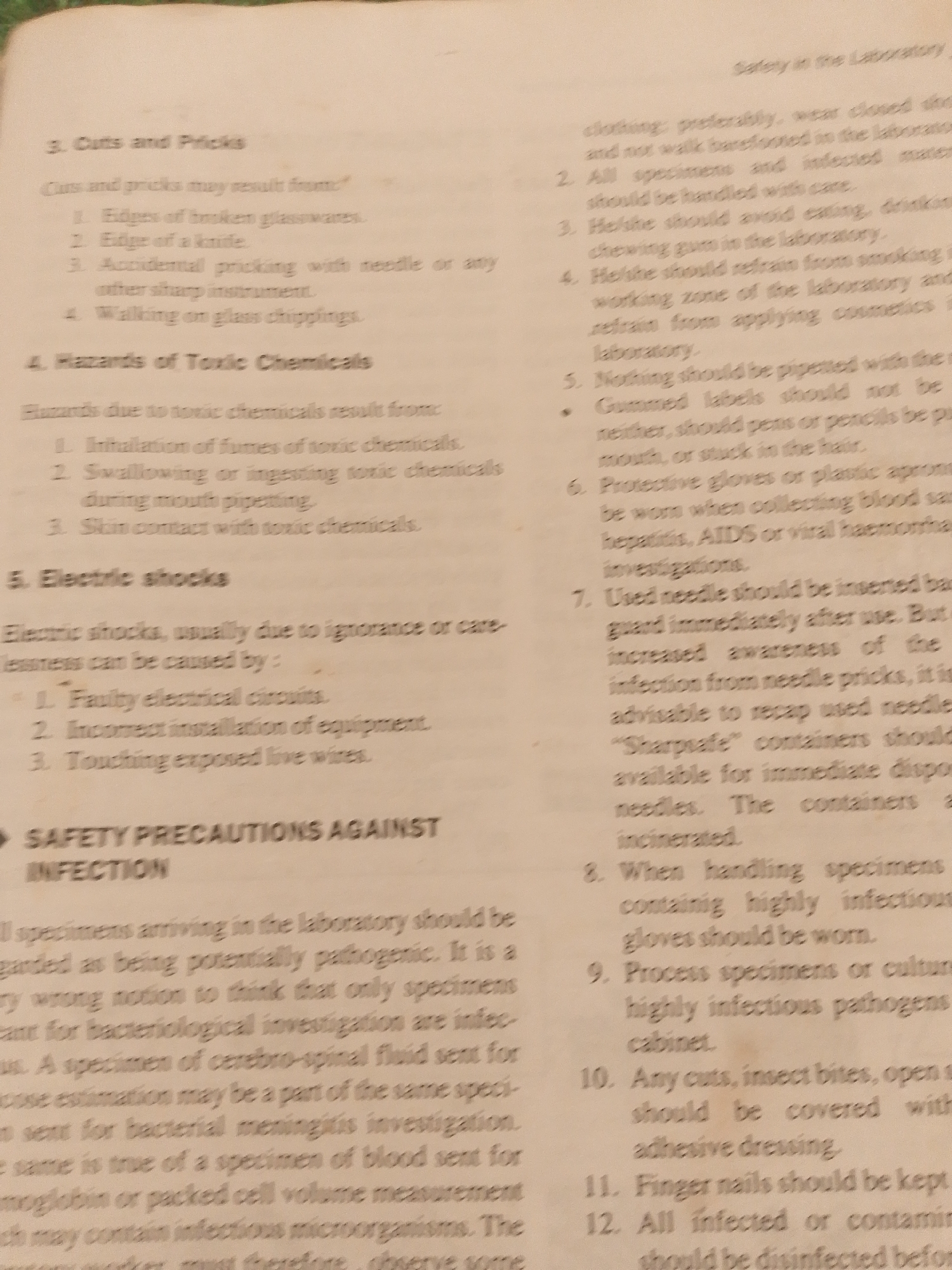 This is a close-up, top-down shot of an open book, likely a manual or guide, filled with text. The pages are slightly creased and show signs of age and use, with some yellowing. The lighting is adequate, allowing for clear readability of the printed words.
The text discusses safety precautions and hazards in a laboratory setting. Specific topics covered include "Cuts and Pricks," "Hazards of Toxic Chemicals," "Electric Shocks," and "Safety Precautions Against Infection." The content is instructional, detailing potential dangers and preventative measures related to handling equipment, chemicals, and biological specimens. There are numbered lists and bullet points that outline specific rules and guidelines, such as avoiding eating or smoking in the laboratory, proper disposal of needles, and the importance of wearing protective gear. The text also mentions specific medical conditions like hepatitis and AIDS, indicating a context related to healthcare or medical research.
Visible text includes section titles like "3. Cuts and Pricks," "4. Hazards of Toxic Chemicals," "5. Electric Shocks," and "SAFETY PRECAUTIONS AGAINST INFECTION." Further details within the text refer to "broken glassware," "needle," "toxic chemicals," "electrical circuits," "exposed live wires," "specimens," "cerebro-spinal fluid," "bacterial meningitis," "blood," "infectious microorganisms," and the use of "protective gloves" and "plastic aprons." The mention of "laboratory" and "specimens" strongly suggests the setting is a scientific or medical laboratory. There are no people, animals, or specific environmental cues visible in the image. The time of day and weather are not discernible. The location context of Jalingo, Nigeria, is not directly evident from the image itself but is provided as external information.