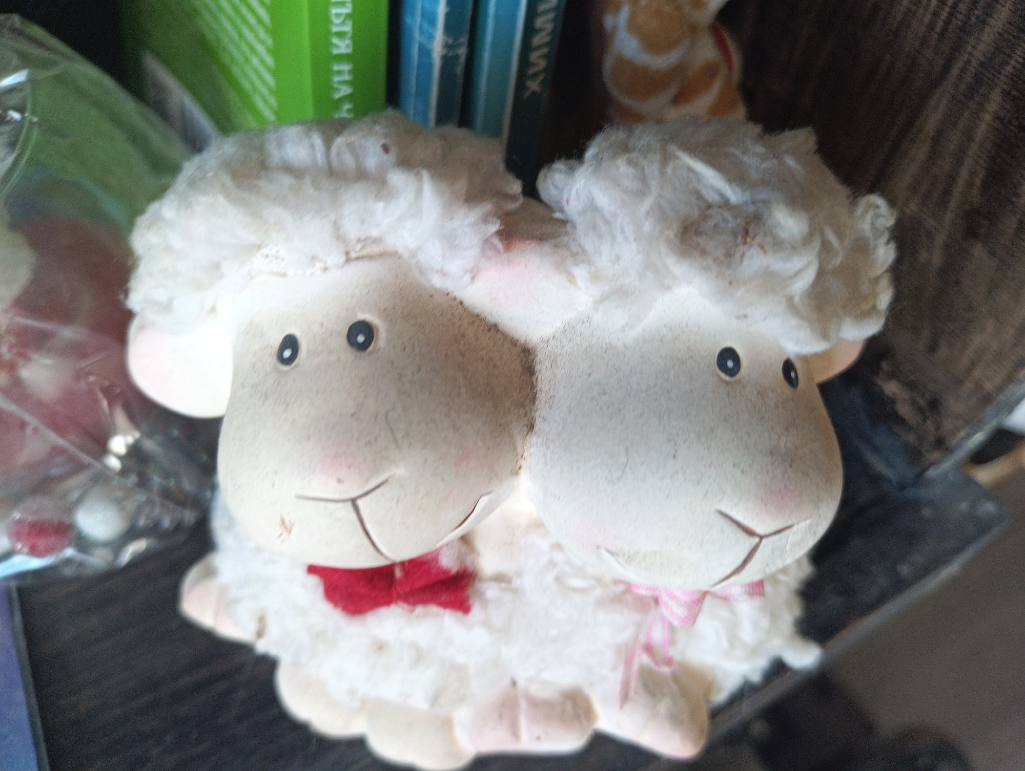 The image shows a close-up of two decorative sheep figurines, likely made of ceramic or resin, with fluffy white wool exteriors. They are positioned side-by-side, facing forward. Each sheep has two black oval eyes and a smiling mouth represented by a simple line. The sheep on the left wears a red bow tie, and the sheep on the right wears a pink bow. Their bodies are rounded and they appear to be sitting. The background is blurred, but suggests a shelf or table with books and possibly other decorative items. The setting does not provide any specific indicators of Karnobat, Bulgaria.