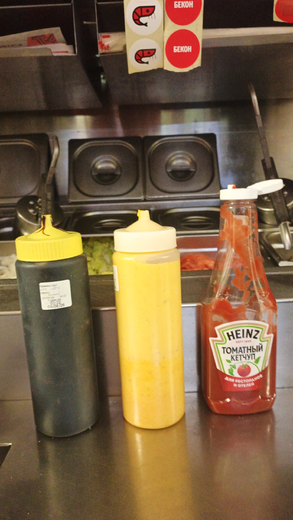 The image shows a close-up of a metal counter with three condiment bottles and two round stickers on a shelf above. From left to right, the bottles are a black squeeze bottle with a yellow nozzle, a tall yellow squeeze bottle with a white nozzle, and a clear bottle of Heinz ketchup with a white lid. The black bottle has a white label with Cyrillic text. The yellow bottle has a white label. The Heinz bottle has a red label with white text and a picture of a tomato. The stickers on the shelf above are round and red, one with a stylized depiction of a shrimp and Cyrillic text that reads "БЕКОН" (BECON, meaning bacon), and another with the same text on a red background. The background appears to be a commercial kitchen or food service area, with stainless steel containers and other equipment visible. There are no people in the image. The scene is lit from above, casting a reflection on the metal counter. The location is a commercial kitchen or restaurant, possibly in Pavlovsky Posad, Russia, given the Cyrillic labeling.