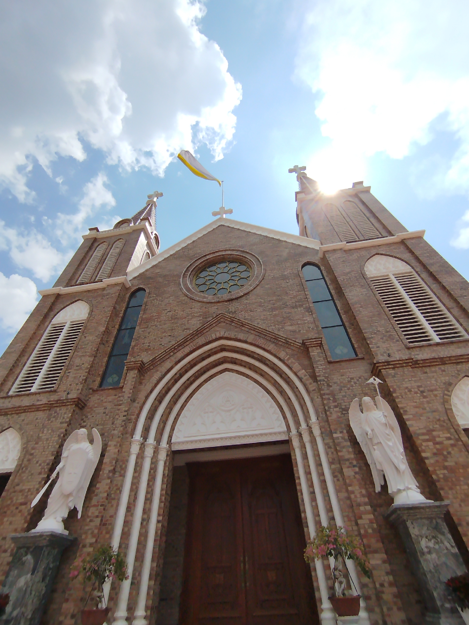 A low-angle shot reveals the façade of a brick church set against a sky with scattered clouds. The church has two towers topped with crosses, with a white and yellow flag flying atop the central structure. The sun shines brightly, creating a lens flare effect.
The church entrance features an arched entryway flanked by two white angel statues holding swords or spears. Small potted plants are placed near the statues.
The building’s architectural style is characterized by the use of brown bricks, tall rectangular windows, and louvred openings. The church is located in Cam Ly - Đà Lạt, Vietnam.