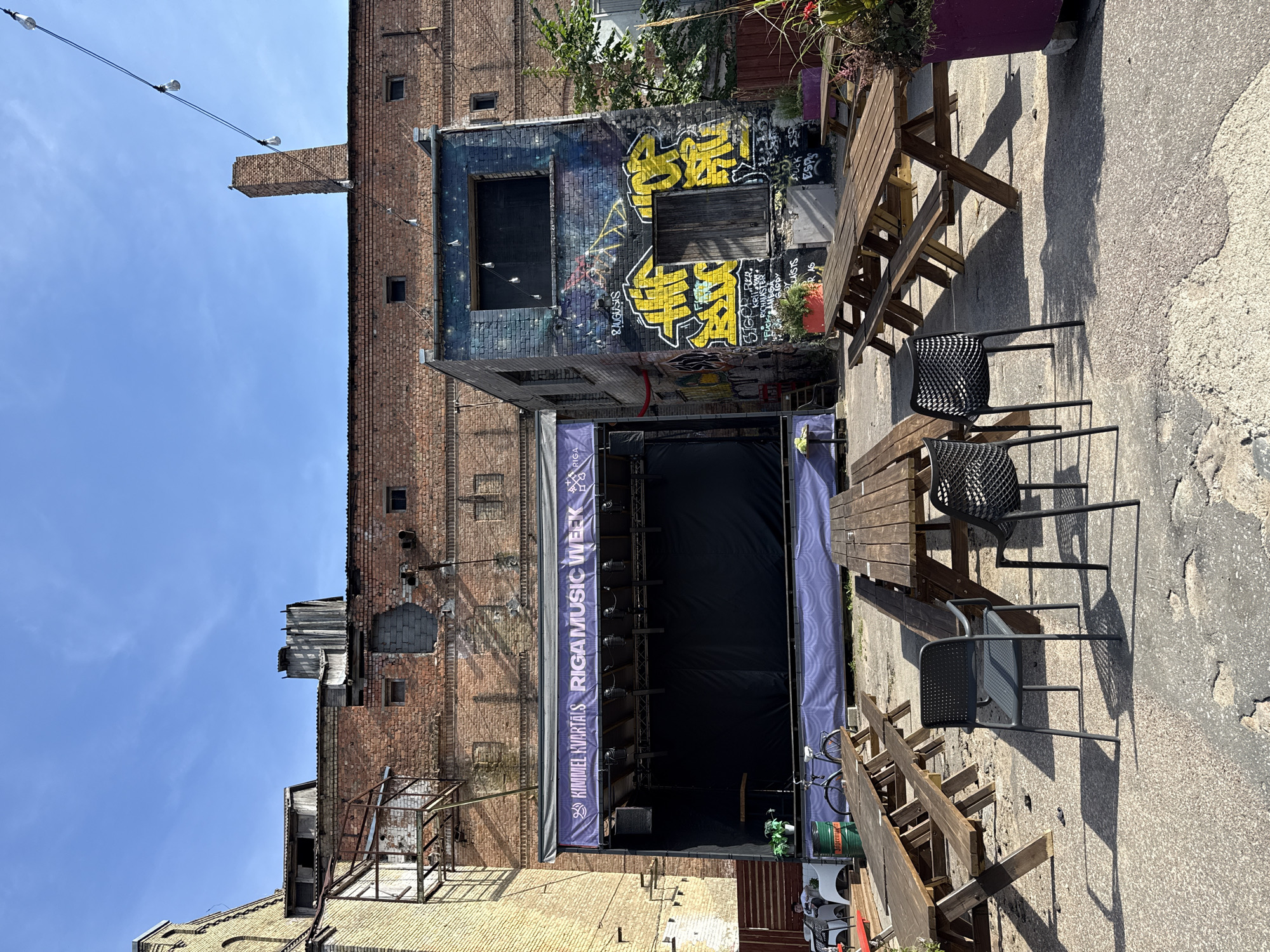 The image depicts an outdoor courtyard in Riga, Latvia, possibly an event space or a beer garden, with a stage set up against a weathered brick building. The stage has a purple banner that reads "KIMMEL KVARTALS RIGA MUSIC WEEK." In front of the stage, there are several empty wooden picnic tables with benches. To the right of the stage, a smaller building is covered in colorful graffiti. String lights hang across the scene. In the foreground, two dark-colored chairs are placed near the picnic tables, also empty. The ground is paved with rough concrete. The overall impression is of a somewhat run-down but functional urban space prepared for an event.