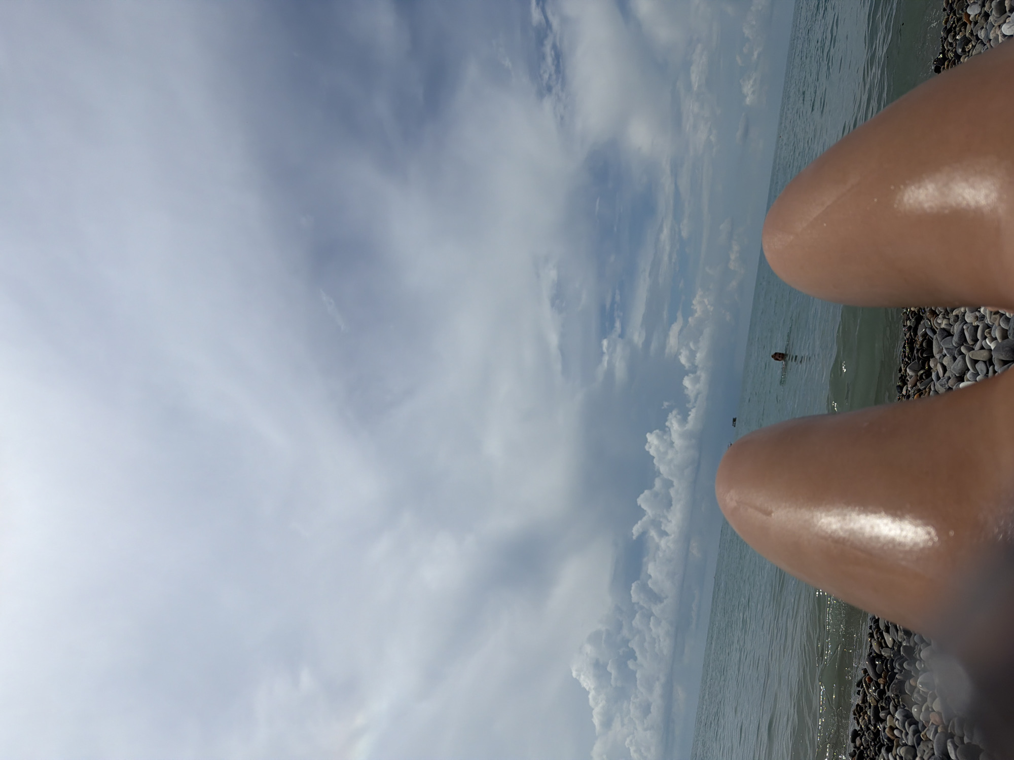 The image captures a first-person perspective of a person lying on a pebble beach, facing the sea. The foreground is dominated by the person's legs, which are shiny, suggesting they are wet or oiled. The beach is covered in smooth, multi-colored pebbles. The sea is calm and turquoise, with small waves lapping at the shore. In the distance, two figures are visible in the water; one appears to be swimming or wading, while the other is further out and less distinct. The sky is filled with dramatic, heavy clouds, interspersed with patches of blue sky. The overall scene depicts a typical day at the beach, likely during the summer months. The location is identified as Sochi, Russia.
