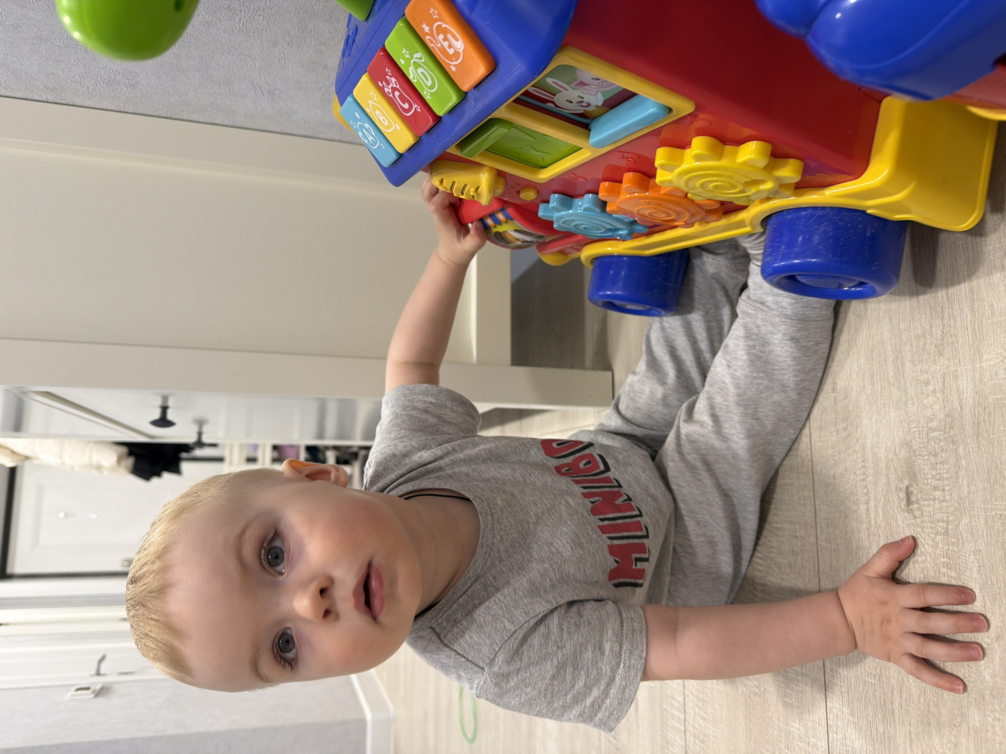 A young, blond-haired child wearing a gray long-sleeved shirt and gray pants is on the floor, appearing to be playing with a toy activity center. The activity center is red, blue, yellow, and green and has colorful buttons and gears. The child is lying on their stomach with one arm reaching up to grasp the toy. The location appears to be inside a building in the city of Votkinsk, Russia, based on metadata. The background includes a white wall, a door, and a light-colored wooden floor.