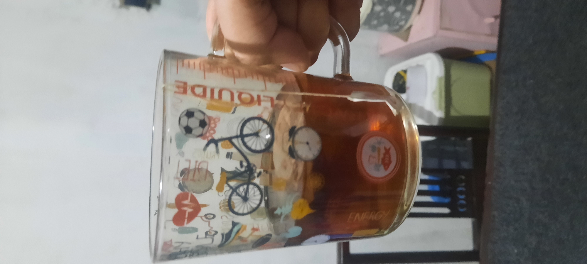 A hand holds a clear glass mug filled with tea, a tea bag steeping inside. The mug is decorated with various small graphics, including a bicycle, a soccer ball, and a heart with an electrocardiogram. The background is blurred, showing a white wall and some furniture, including a green plastic bin. The scene appears to be indoors, possibly in a home or office. No specific location in North Jakarta, Indonesia, is visible.
