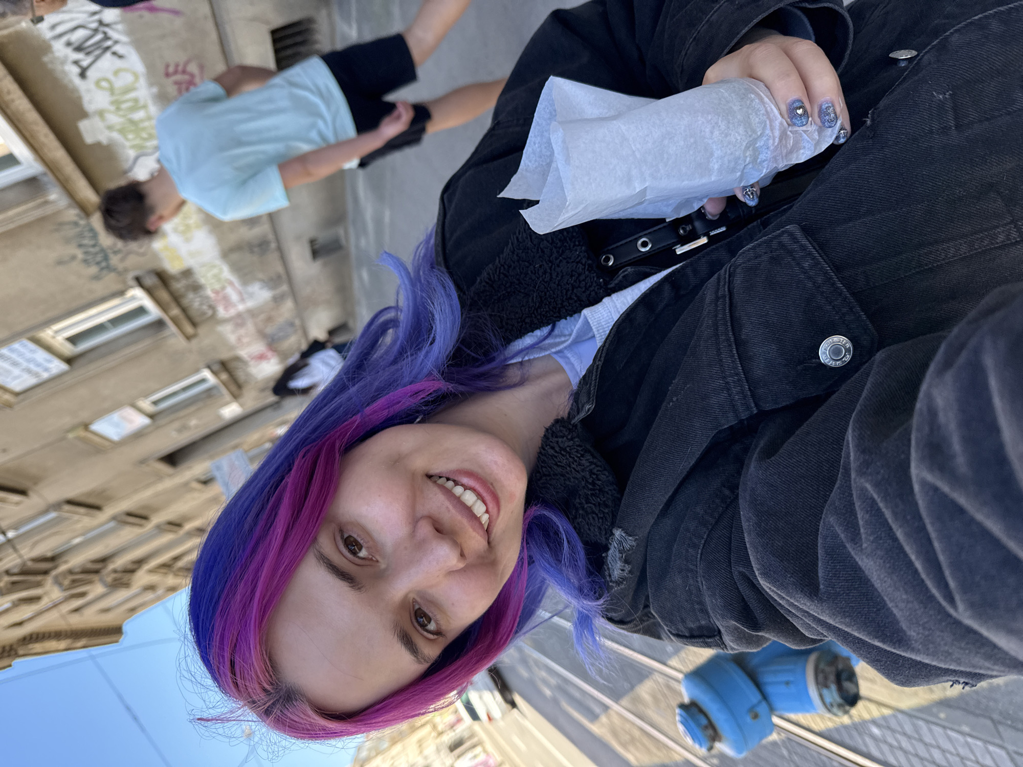 A medium, angled shot captures a person with dyed purple and pink hair, smiling at the camera while holding a paper-wrapped item, possibly food. The person is wearing a black denim jacket with a shearling collar. In the background, a second person in a light blue shirt and dark shorts is partially visible. The setting appears to be a street in City of Zagreb, Croatia. Graffiti is present on the walls of the buildings in the background. A blue fire hydrant is visible near the bottom of the frame.
