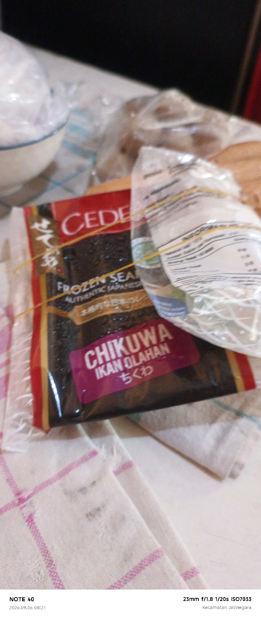 A package of "CEDEFROZEN SEAN AUTHENTIC JAPANESE CHIKUWA IKAN OLAHAN" is on a white cloth with pink stripes. The package is secured with a yellow rubber band. The product appears to be food. The lower right corner of the image contains text indicating "Kecamatan Jatinegara," which is a district in East Jakarta, Indonesia. The timestamp "2026.09.06 08:21" and camera settings "23mm f/1.8 1/20s ISO7033" are also visible.
