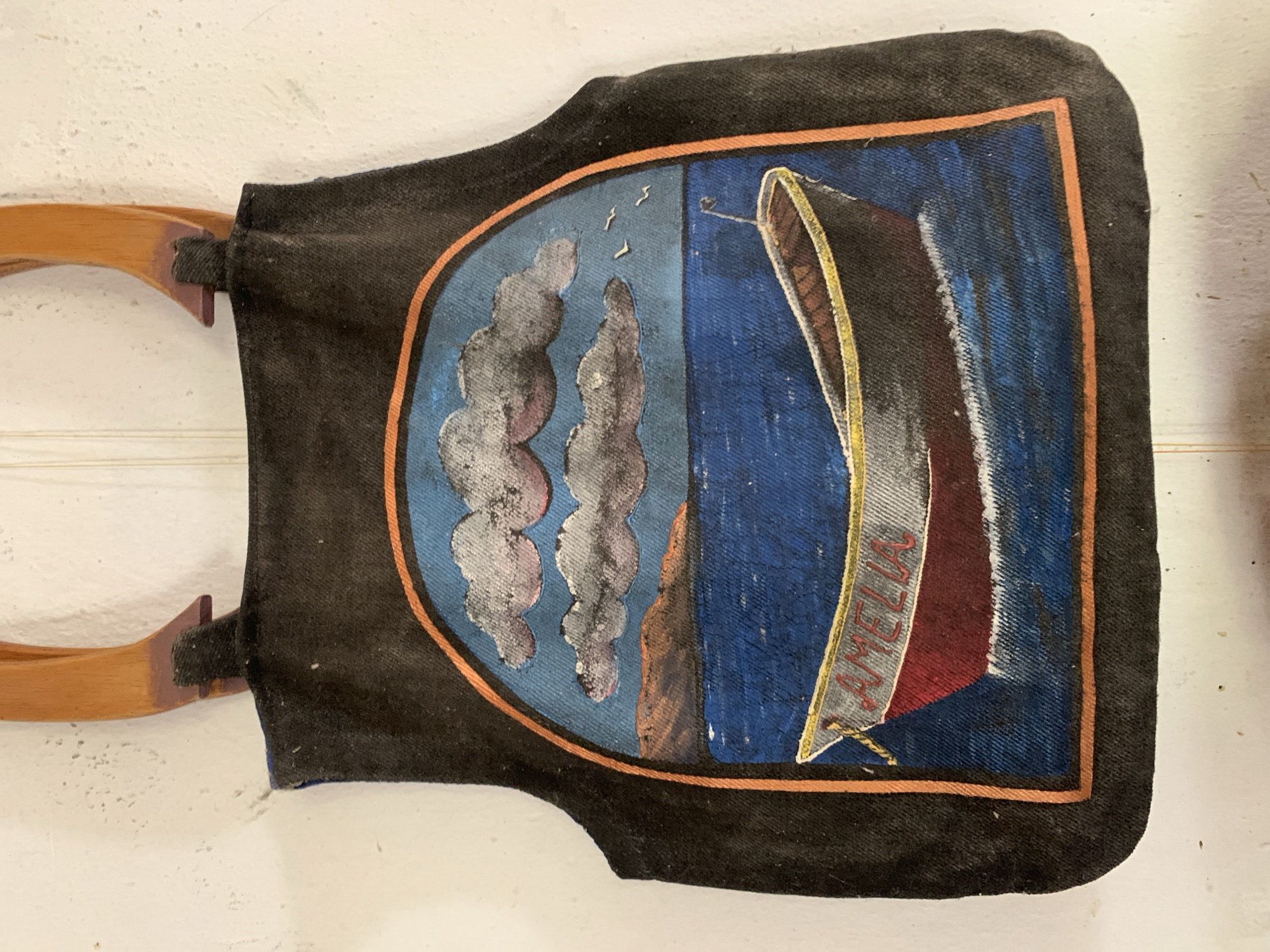 A rectangular, black fabric object with a painted design is displayed against a white, textured surface. The object, suspended by two wooden rods, features a central shield-shaped design outlined with a thin orange border. The painted scene depicts a maritime theme with blue background suggestive of water, the name "AMELIA" is written on a ship’s sail. To the left of the ship, there are elements resembling stylized grey clouds, a rocky shoreline, and three small, bird-like shapes in the sky. The paint has a faded quality. The object appears worn and potentially vintage. The location cannot be inferred from this image.