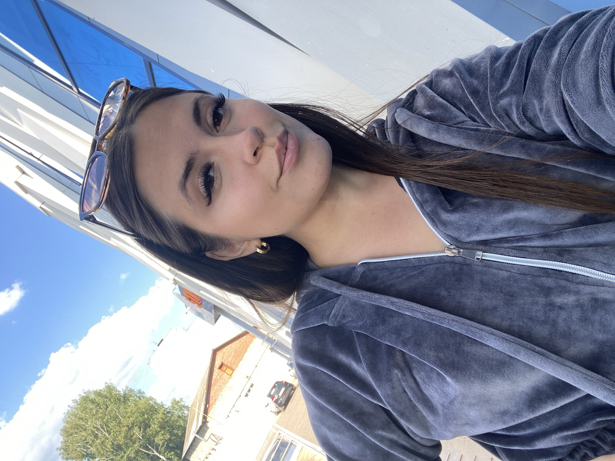 In Chaikovsky, Russia, a young woman with long brown hair and false eyelashes poses for a selfie outdoors. She is wearing a dark gray velour zip-up hoodie and gold hoop earrings. Sunglasses are perched on her head. Behind her, a modern building with reflective blue windows and white architectural elements stands tall against a bright blue sky dotted with white clouds. To the left, a brick building with an orange window is visible, and a parked dark car can be seen on a road. A leafy tree is also present in the background. The woman is looking directly at the camera with a slight smile.