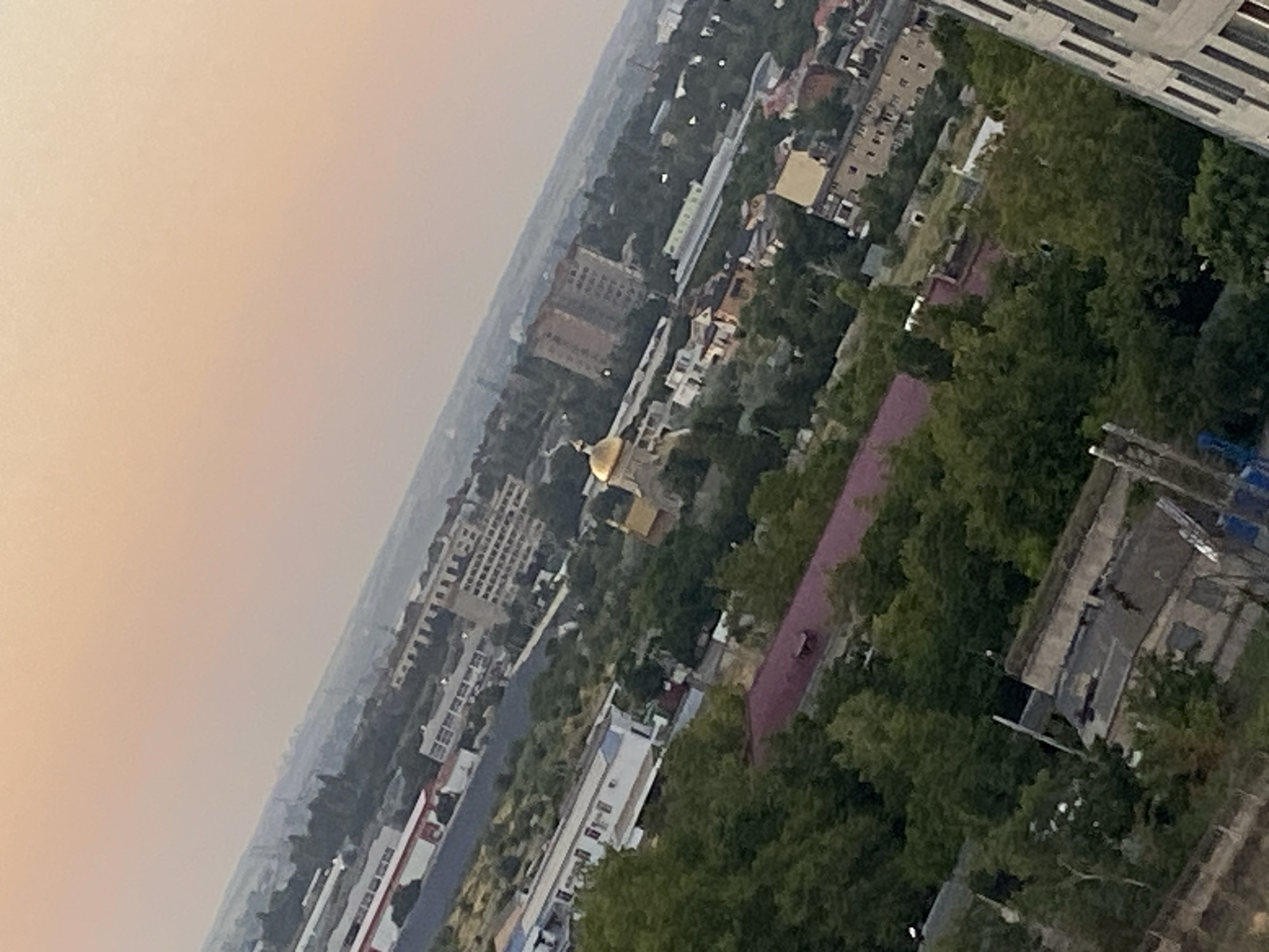 The image captures a tilted, aerial view of a city at dusk, with a hazy, orange sky dominating the upper portion. The landscape is a mix of urban development and greenery, with buildings of varying sizes and architectural styles interspersed with dense trees. In the middle ground, a golden-domed building, likely a church, stands out against the surrounding structures. Several multi-story apartment buildings are visible, as are smaller residential houses and industrial-looking facilities with long, low-rise buildings. The presence of trees suggests a city that incorporates green spaces. The overall impression is a moderately sized city with a mix of older and newer construction, set against a backdrop of an atmospheric sunset. Based on the provided crop hints indicating it is of Chornomorsk, Ukraine, the scene depicts a section of that city during twilight.