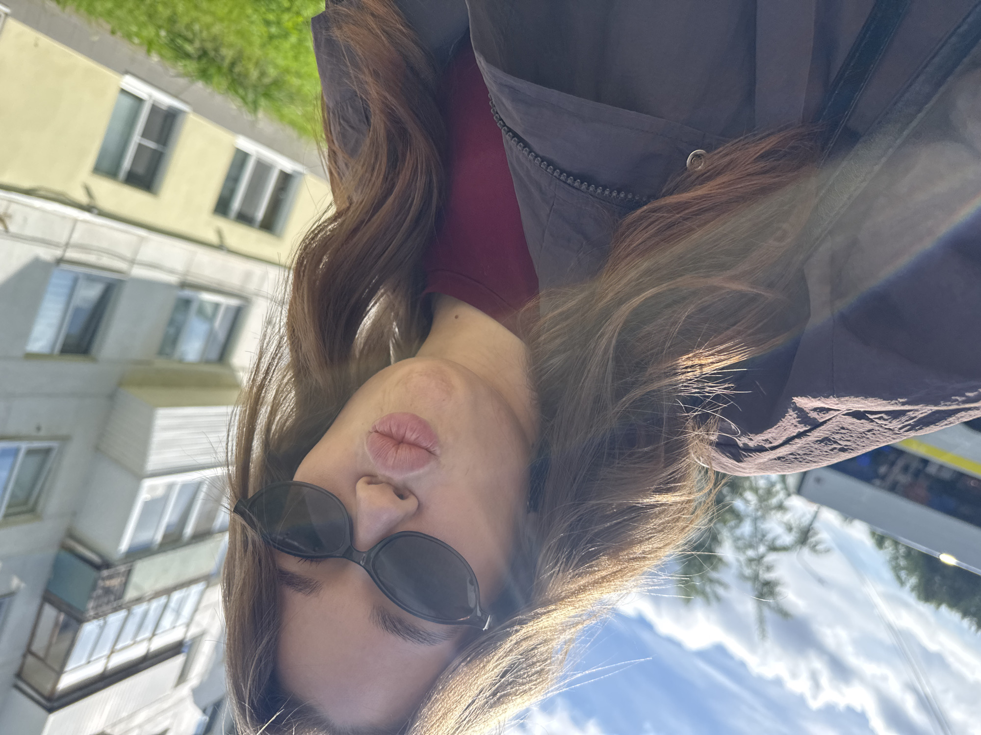 A young woman with long brown hair and sunglasses makes a pouty face for a selfie. She is wearing a maroon t-shirt and a dark jacket. In the background, a light yellow apartment building with numerous windows is visible. The sky is blue with white clouds. The scene appears to be outdoors, possibly on a street in the city of Chaikovsky, Russia, given the architectural style of the building.