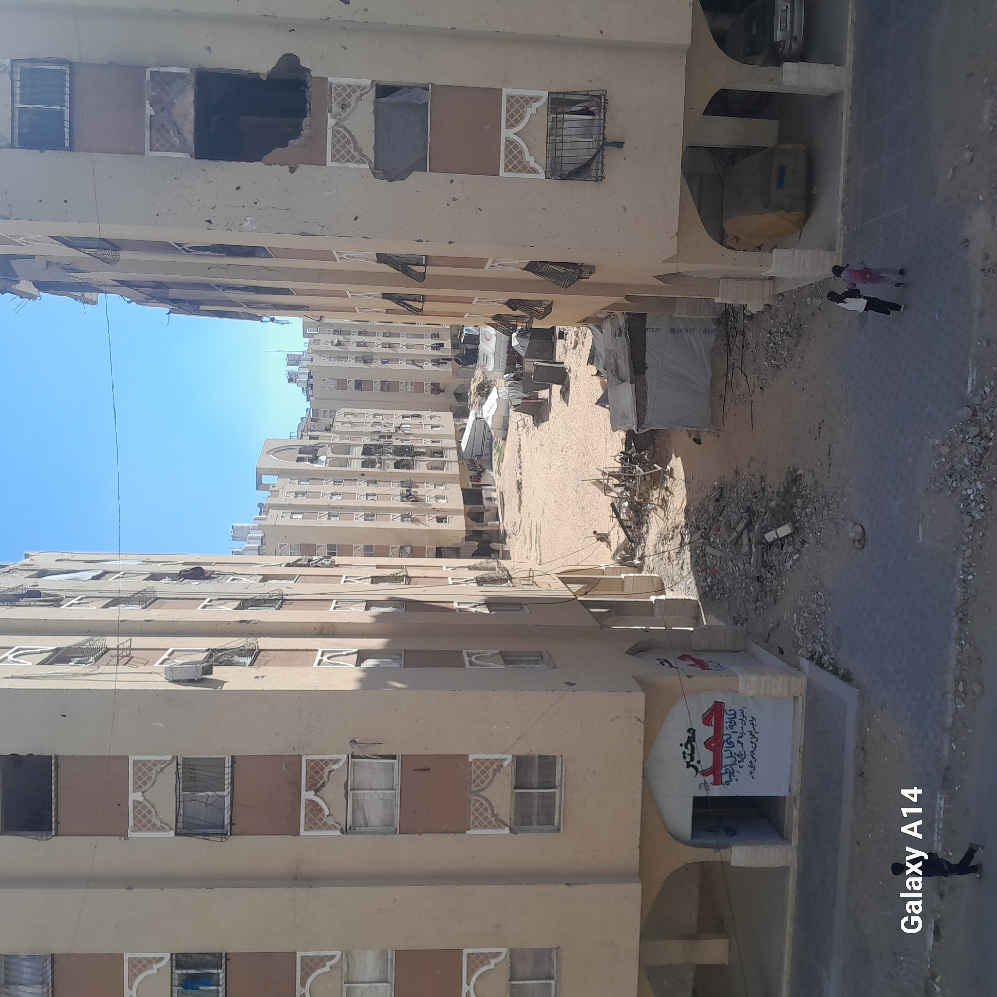 The image depicts a street scene in an urban environment, likely the city of Unknown, Unknown. Multi-story, beige buildings line both sides of a sandy central area with visible damage, possibly due to conflict or disaster. Some buildings show signs of disrepair, with missing or damaged windows. Several makeshift shelters or tents are visible in the sandy area, suggesting a displacement of people. Debris and rubble are scattered on the ground. Two people walk along the paved road adjacent to the sandy area, and a person walks away from a building with a sign written in Arabic, possibly indicating a medical facility. The sky is clear and blue. A "Galaxy A14" watermark is visible at the bottom right corner of the image.