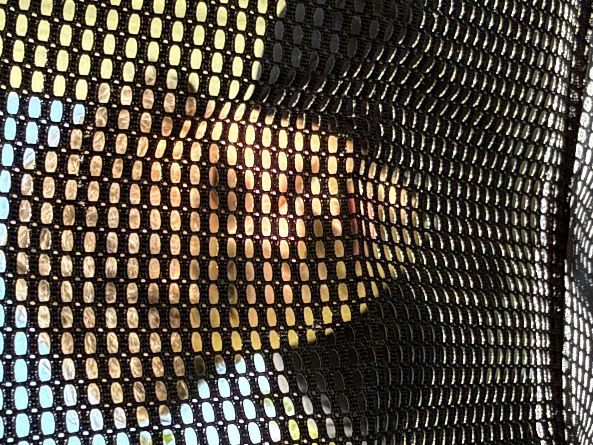 The image depicts a close-up view of a black mesh fabric with a grid-like pattern. Light filters through the mesh, creating a varying color gradient from blue and yellow to a warm, orange hue. In the center of the image, an indistinct human face appears, partially obscured by the mesh, with light highlighting the features. The overall effect suggests someone looking through a screen or barrier. The location in Bar, Montenegro, is not explicitly evident from the visual content alone; this information would be inferred or based on context outside the image.