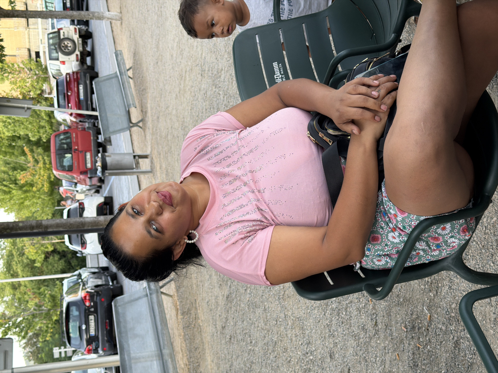 The image depicts a woman sitting on a dark green plastic chair, outdoors. She is wearing a pink t-shirt adorned with sparkling embellishments and a floral skirt. Her left arm is resting on her lap, and her hands are clasped. Behind her, a young child in a white t-shirt is partially visible, also seated. The background reveals a parking lot with several cars, including a black SUV and a red van, and trees. The ground is covered with gravel. The scene appears to be located in Hostalric, Spain.
