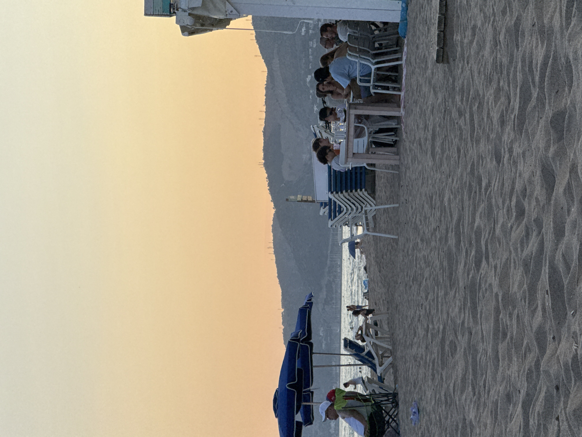 This image depicts a beach scene in Castelldefels, Spain, during what appears to be late afternoon or early evening, given the soft, warm hues of the sky. The setting is a sandy beach with the gentle waves of the Mediterranean Sea visible in the background.
The primary subjects are a group of people gathered at tables set up on the sand, likely at a beachfront restaurant or cafe. They are seated, engaged in conversation, and appear to be enjoying a meal or drinks. The people are diverse, with individuals of various ages and genders. Some are dressed casually, in t-shirts and shorts, fitting for a beach environment.
Scattered around the scene are typical beach objects. There are numerous white lounge chairs, many stacked neatly, suggesting they belong to the establishment or are available for rent. Several beach umbrellas, including a prominent large blue one, provide shade. The sand itself is a textured element, showing the characteristic ripples left by the wind or water. In the distance, a mountainous landscape rises, providing a backdrop to the coastal setting.
The activity taking place is leisurely. People are relaxing, socializing, and dining by the sea. The overall mood seems tranquil and pleasant, indicative of a typical vacation or leisure day at the beach. The time of day is suggested by the warm, soft light illuminating the sky, casting a gentle glow over the scene. There are no visible text elements in the image.