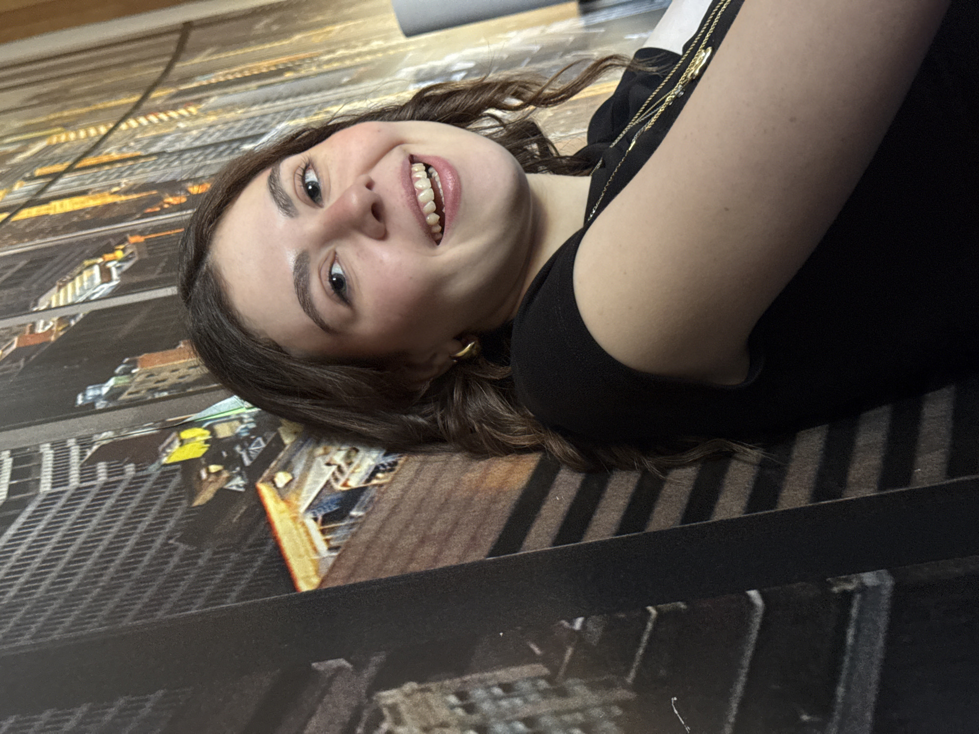 A young woman with light skin and brown hair is lying on her side on a surface that appears to be a printed image of a city landscape, possibly New York City, as skyscrapers and grid-like patterns are visible. She is wearing a black top and gold jewelry. She is smiling broadly, facing the camera. The location is not Mirandela, Portugal, but rather a setting containing an image of New York City.