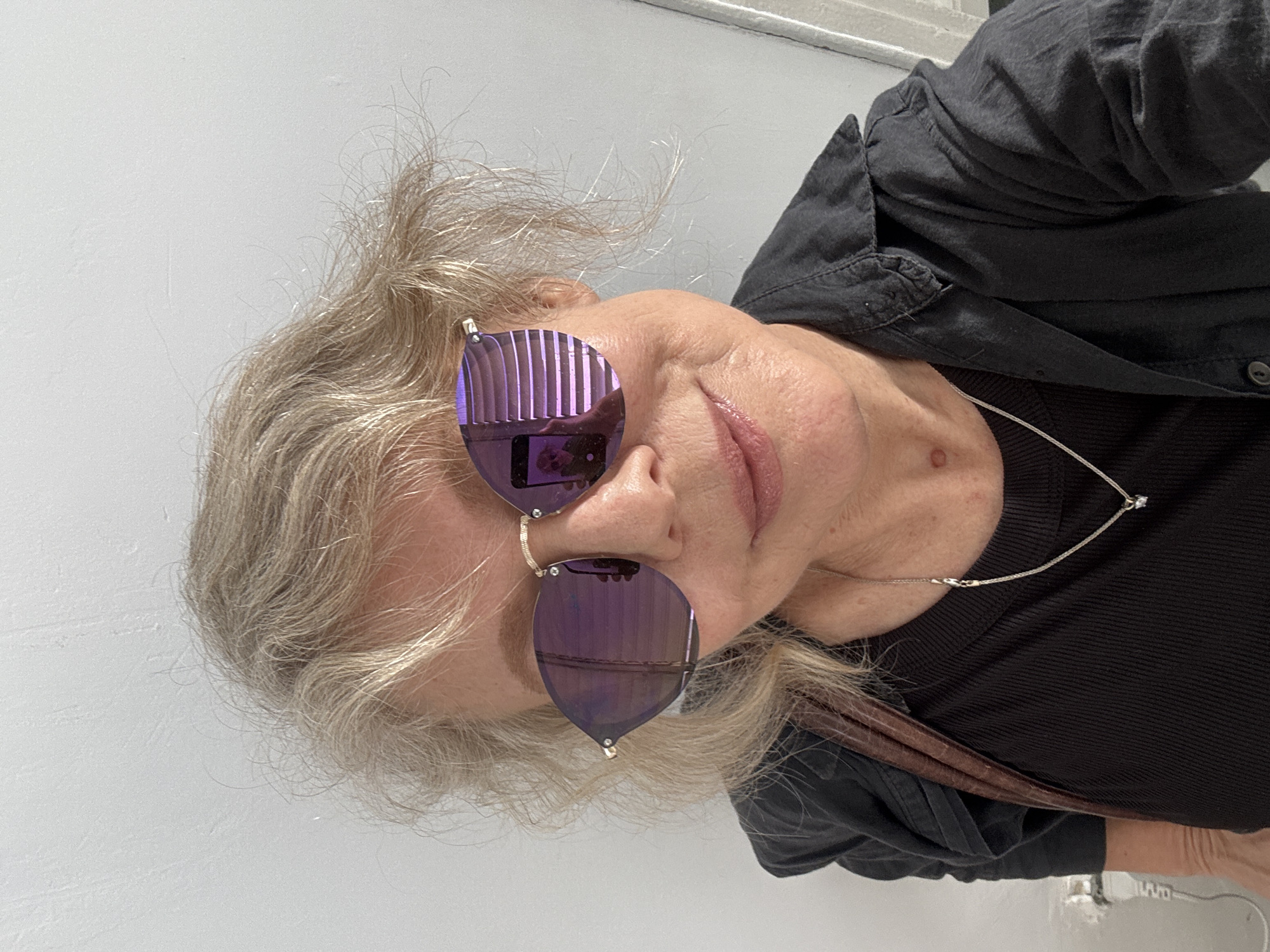 A selfie depicts a mature Caucasian woman, with a medium complexion, wearing distinctive purple sunglasses with a vertically ribbed pattern. Her hair is grey and styled casually. She is wearing a black top and a silver necklace. The background appears to be a plain white wall. There is no indication that the image was taken in Ramat Gan, Israel.
