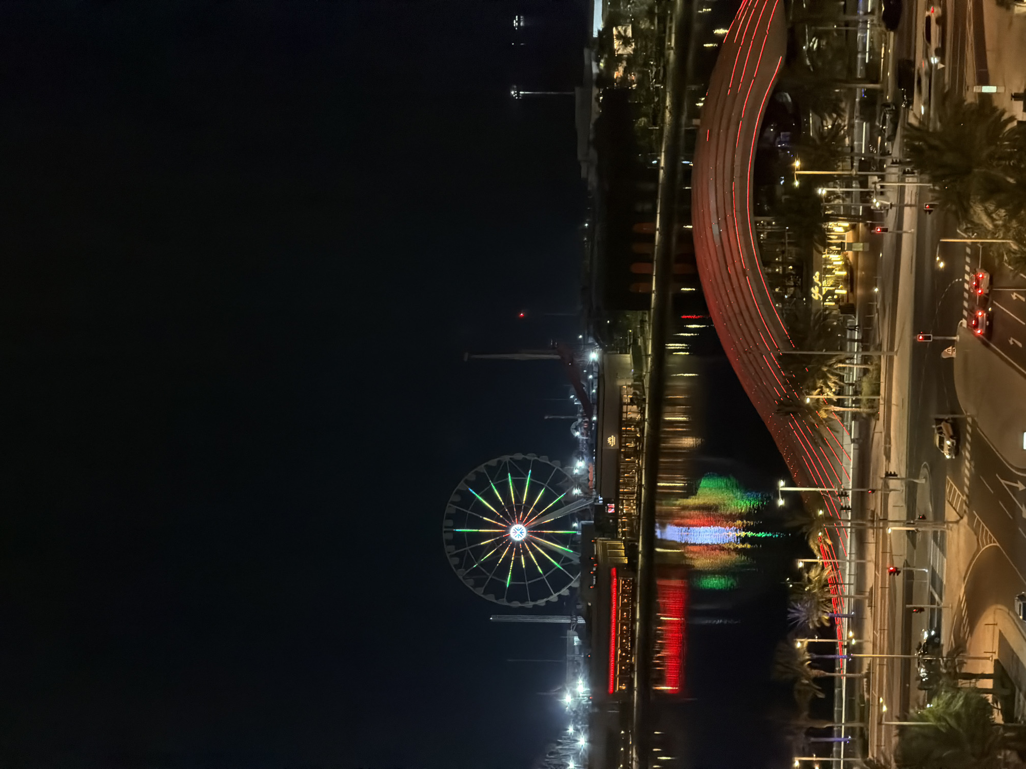 The image captures a nighttime scene in an urban waterfront area. The prominent subjects are a Ferris wheel illuminated with colorful lights and a bridge with red lights outlining its structure. The Ferris wheel, with its spokes radiating from the center, displays a vibrant pattern of green, yellow, and blue lights. The bridge curves gracefully over the water, its red lights reflecting on the dark surface.
The scene is set by a body of water, likely a river or canal, that separates the illuminated structures from a street on the right side of the image. On the street, traffic lights are visible, and several cars are on the road, suggesting the time is late evening or night. The presence of palm trees along the waterfront and road hints at a warm climate or a landscaped urban environment. The overall atmosphere is one of a lively and brightly lit cityscape at night.
Notable details include the reflections of the colorful lights on the water, creating a mirrored effect of the illuminated features. The sky is dark, emphasizing the artificial lighting of the city. The activity taking place is essentially the city at night, with transportation and recreational elements like the Ferris wheel active. No people are clearly discernible in the image. There is no visible text in the image.