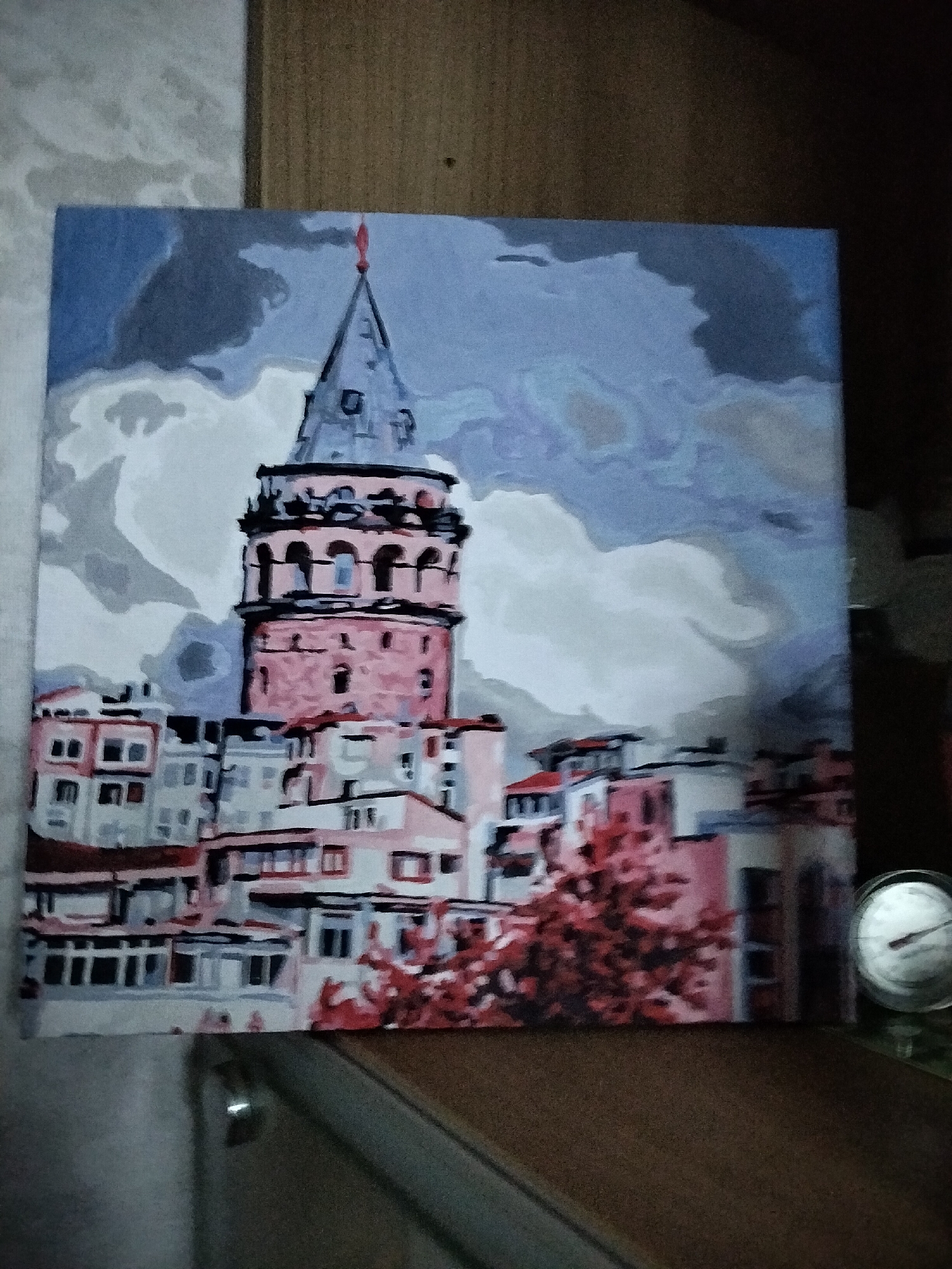 The image displays a painting of the Galata Tower in Istanbul, Turkey. The painting is done in a simplified, almost abstract style with bold blocks of color. The tower itself is depicted in shades of pink and grey, with arched windows and a conical roof topped with a red flag. Surrounding the tower are buildings rendered in similar muted tones of pink, white, and grey. The sky is a dramatic blend of blues, greys, and whites, suggesting cloudy or stormy weather. In the foreground, on the right side of the image, a circular gauge, possibly a thermometer or barometer, is partially visible. The painting is placed on a surface that appears to be a wooden table or desk, with a textured wall or background behind it. The lighting is dim, casting a shadow on the right side of the painting. The location of Nizhnedonskoy, Russia, is not present in this image; the subject matter is clearly Istanbul.