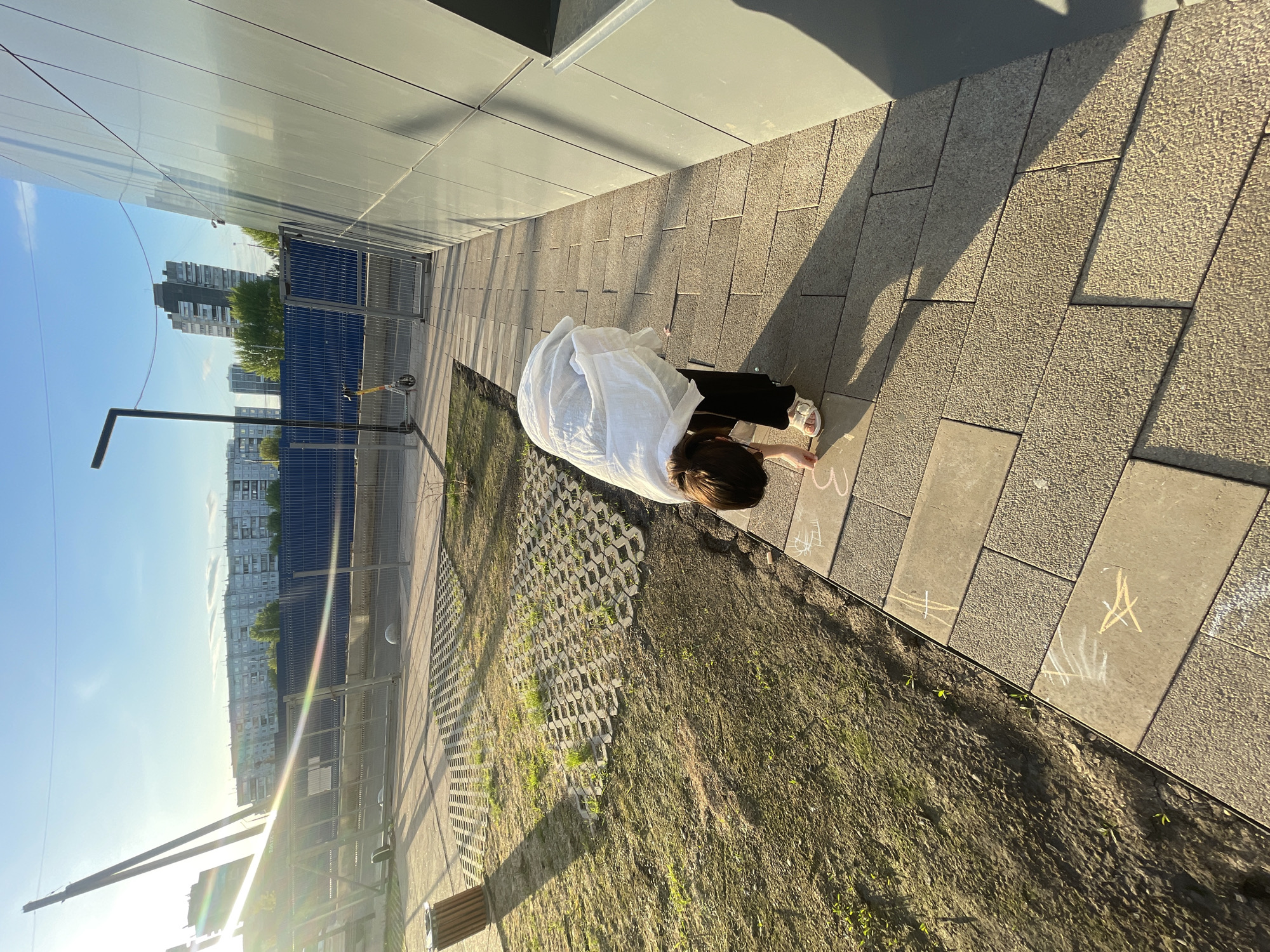 A person is bending over, possibly drawing with chalk on the sidewalk in front of a building in Krasnoyarsk, Russia. The person is wearing a white shirt and dark pants and is facing toward the building. The sidewalk is made of gray rectangular pavers. Next to the sidewalk is a dirt area with grass growing through a honeycomb structure. The building is a light gray color with reflective windows. In the background are apartment buildings under a blue sky. A lamppost is visible to the left. There are white chalk markings on the sidewalk.
