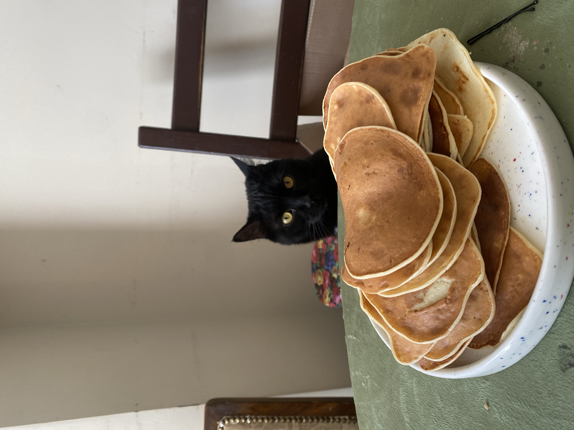 A black cat with yellow eyes peeks out from under a dark brown chair towards a plate of pancakes. The plate is white with a blue speckled pattern and is positioned on a light green surface. A black bobby pin is visible near the edge of the plate. The scene appears to be indoors. The location of Moscow, Russia is not discernible from the image.