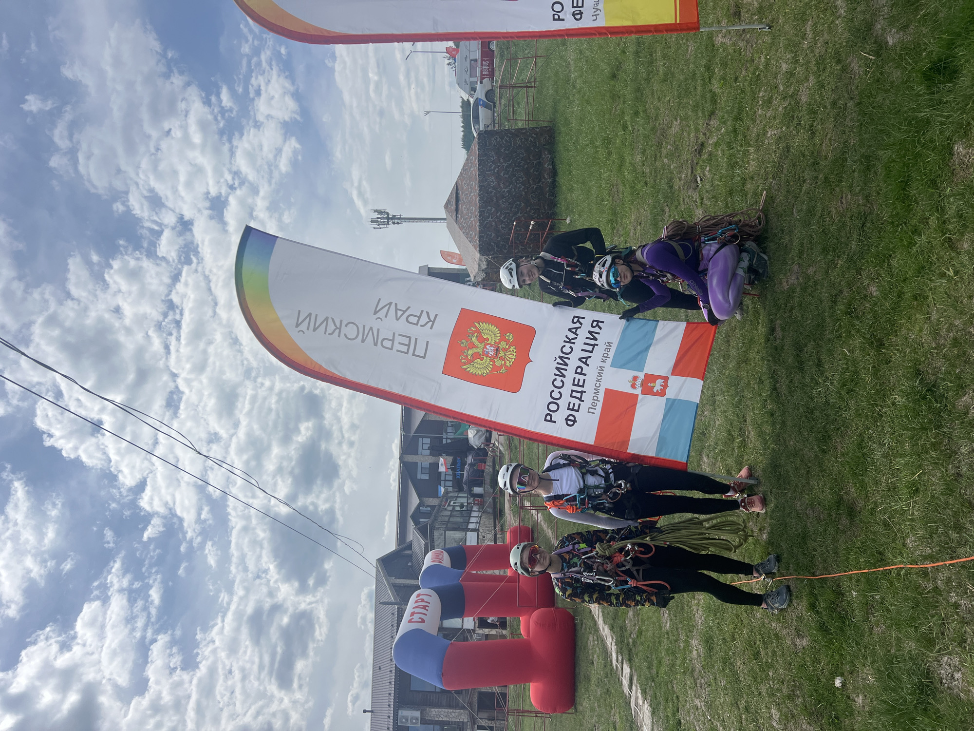The image captures an outdoor event in Khvalynsk, Russia, featuring participants in climbing gear and flags representing the Perm Krai region of the Russian Federation. Two individuals in climbing harnesses, helmets, and goggles stand near a "START" arch in the colors of the Russian flag. Behind them, a white flag bearing the Perm Krai coat of arms and Cyrillic text is prominently displayed. Three other individuals are visible further back on the grass, also wearing climbing gear. The background includes buildings, a cell tower, and a vehicle. The scene is set under a partly cloudy sky.