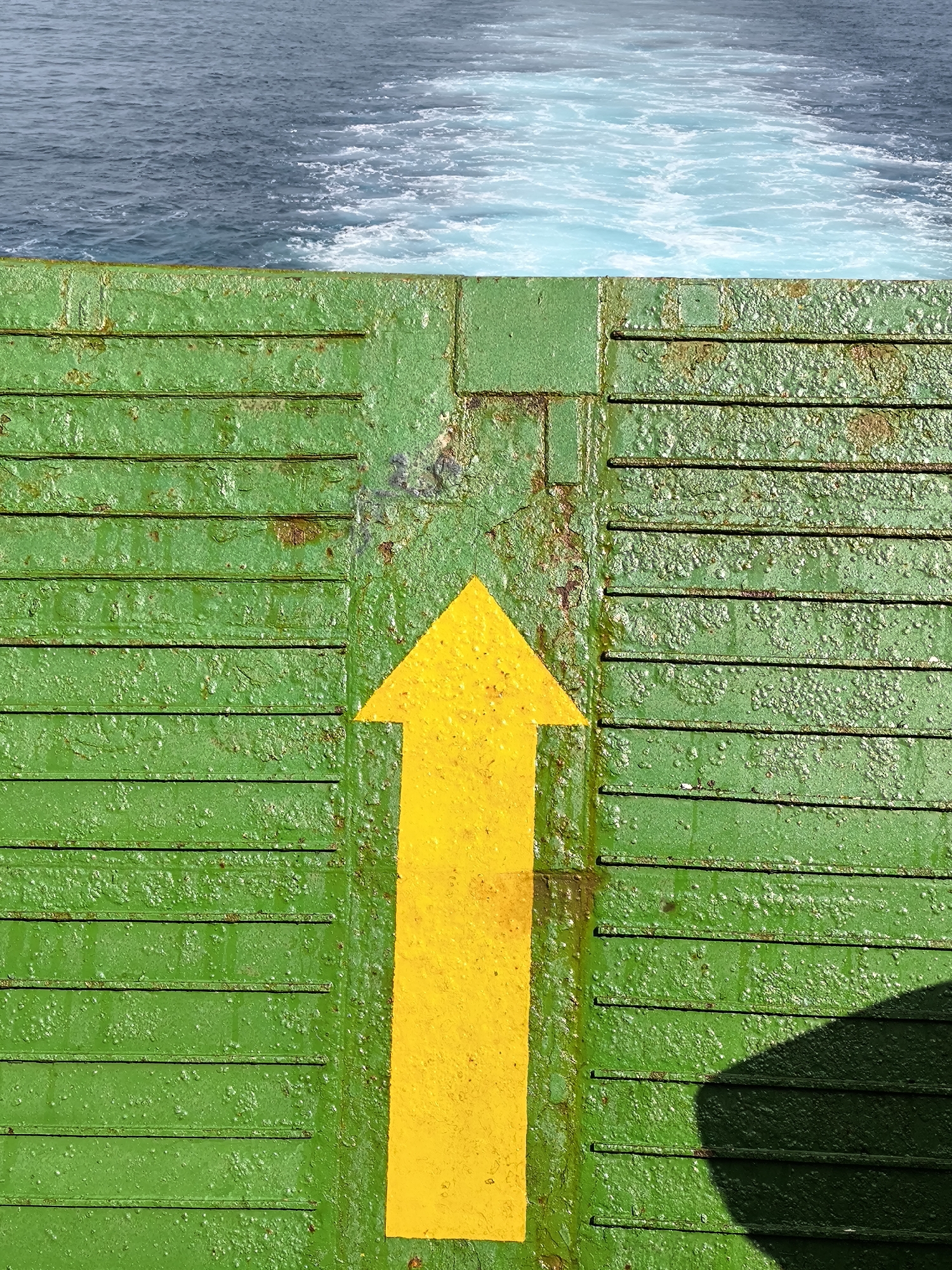 The image captures a vibrant yellow arrow painted onto a heavily textured, weathered green surface, likely the deck or ramp of a marine vessel. The arrow is positioned centrally, pointing directly upwards from the bottom of the frame. The green surface exhibits significant wear and tear, with noticeable rust spots, chipped paint, and an uneven, bubbled texture, indicating long-term exposure to the elements. Horizontal ridges or grooves are visible across this surface, possibly for traction or structural purposes.
In the upper third of the image, a body of water is visible, dominated by a foamy, light blue and white wake that transitions into darker blue, choppier water further back. This wake clearly indicates that the scene is viewed from a moving boat or ferry, looking towards its stern. The bright illumination and a distinct dark shadow cast on the lower right portion of the green surface suggest that the photograph was taken on a clear, sunny day. No people or specific identifying landmarks are present. The only visible "text" is the bright yellow directional arrow.