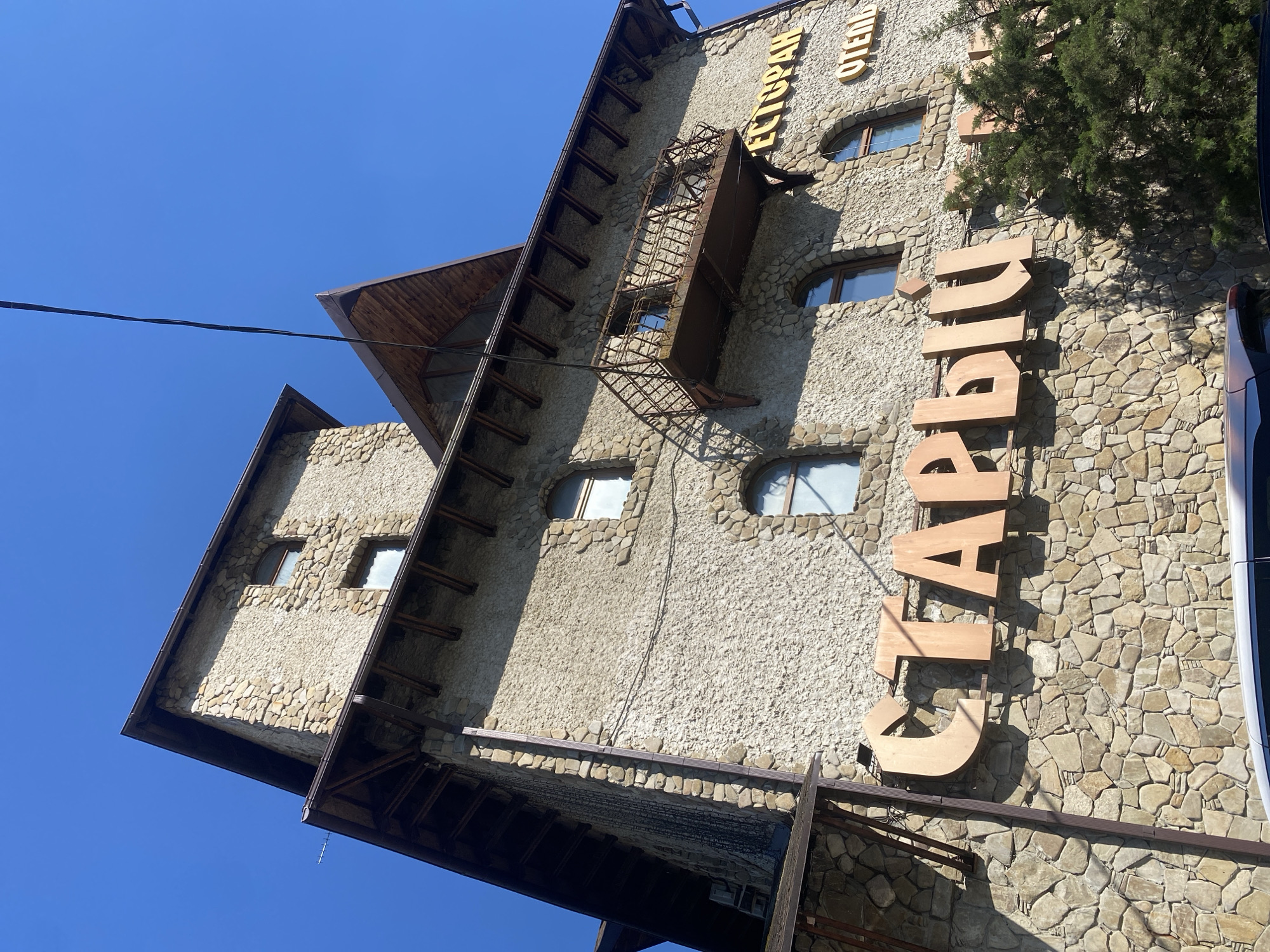 The image shows a stone building with an outdoor restaurant. The building is tall and has a rustic appearance, with many windows and a balcony. There is a sign on the building that reads "СТАРЫЙ" which is Russian for "old". The building is located in the city of Goryachiy Klyuch, Russia. The sky is clear and blue, and there is a tree in the foreground.