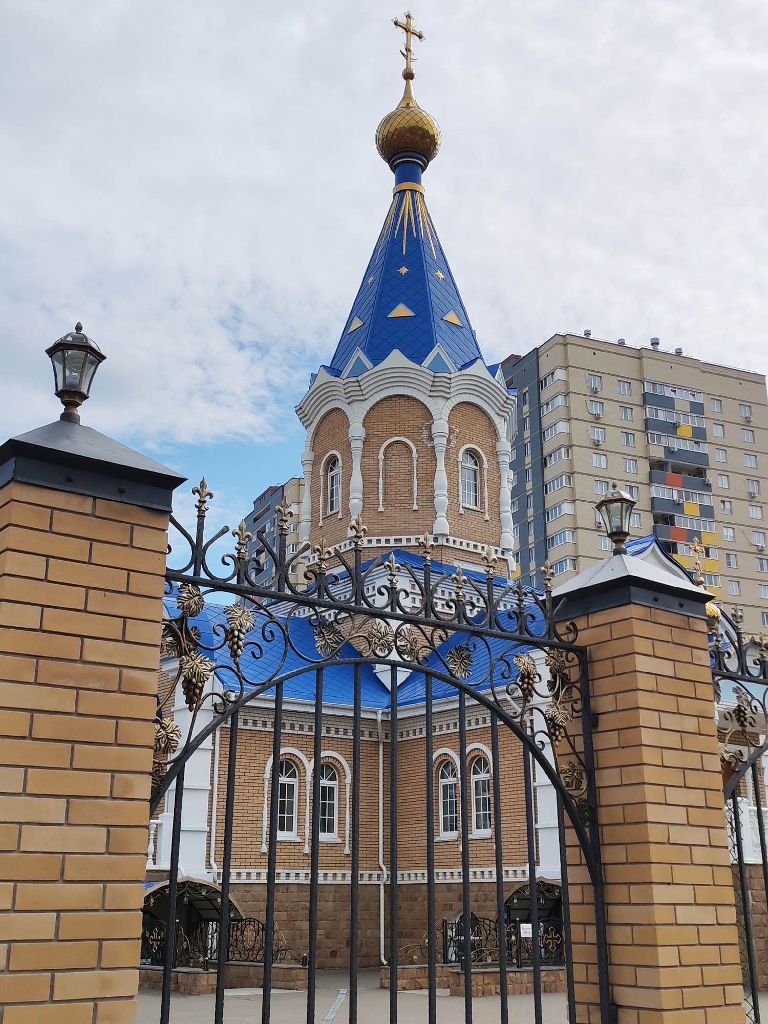 The image depicts the Church of St. Nicholas in Izhevsk, Russia. It is viewed through a wrought-iron fence with brick pillars topped by lamps. The church features a blue-tiled roof with a golden dome and cross. The building's facade is primarily brick with white trim around the windows and architectural details. In the background, a multi-story residential building is visible. The sky is cloudy. No people are visible in the frame.