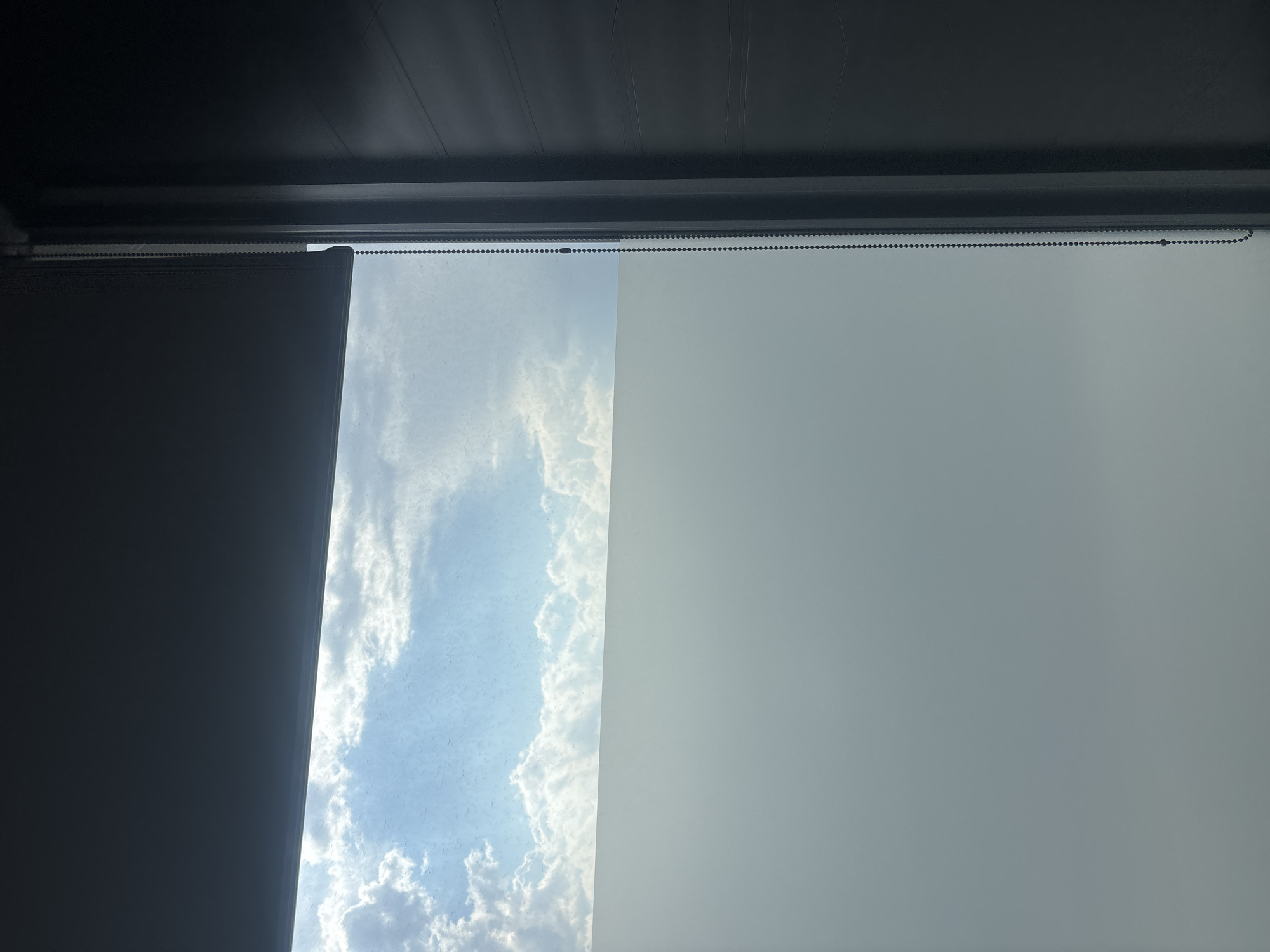 The image shows a window with partially drawn blinds. The left side of the window is covered by a dark gray blind. The center portion reveals a view of a bright blue sky with white clouds. To the right, a lighter gray blind covers the remainder of the window. A beaded chain hangs along the top of the window frame, extending across the length of the blinds. The top of the window frame is dark gray. There is no indication of the location being in Buenavista, Mexico, or any other location. No people, subjects, actions, or interactions are visible in the image.