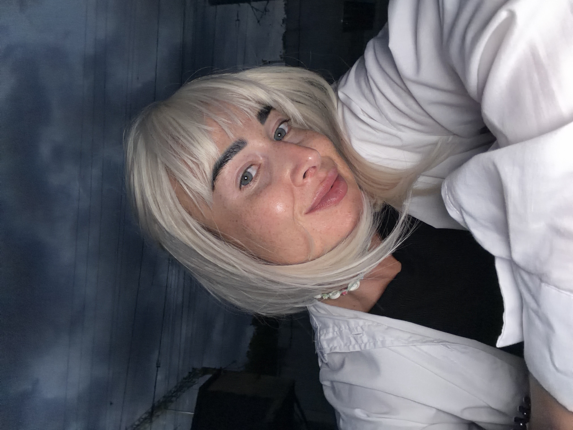 A woman with platinum blonde hair and bangs, wearing a white collared shirt over a black garment and a multi-colored beaded necklace, is captured in a selfie taken outdoors at dusk. Her head is tilted, resting on her arm, and she is looking at the camera with a slight smile. The background features a dark, cloudy sky with visible power lines and indistinct structures, suggesting an urban or suburban environment. The image was taken in Sochi, Russia.