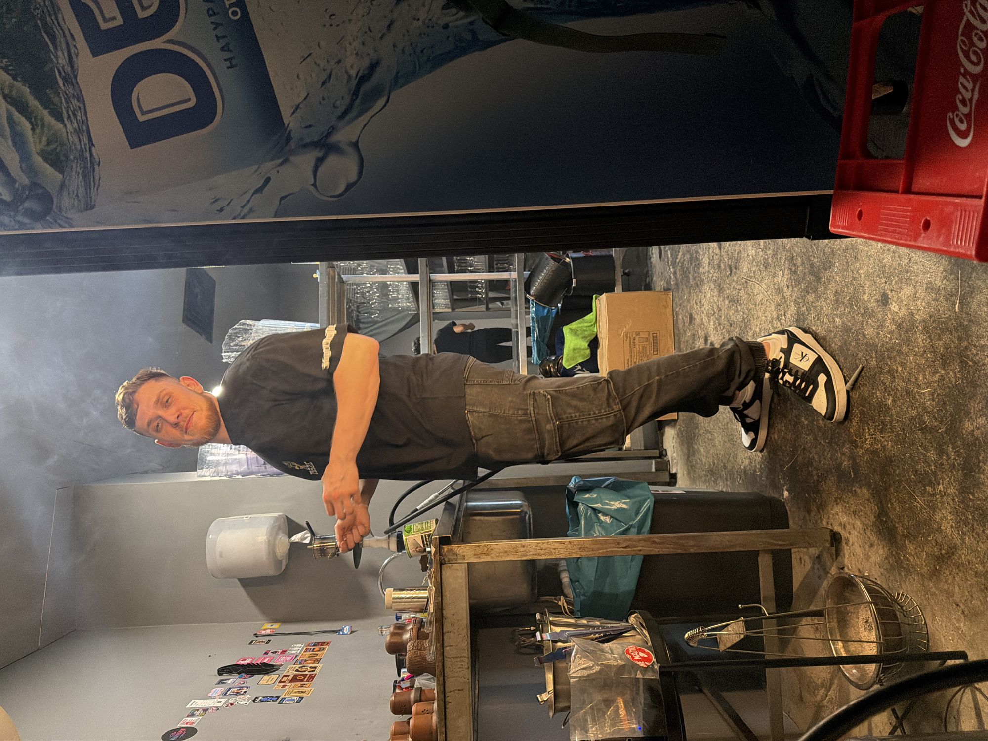 A man is washing something in a metal sink in a room with gray walls. He is wearing a black t-shirt and black pants, and black and white sneakers. There is a paper towel dispenser on the wall, and several stickers. Behind him, there is a counter with items on it. The location is likely a commercial kitchen or prep area. Coca-Cola containers are visible. A water brand DEVIN in Bulgarian is seen. Since DEVIN water is very common in Bulgaria, and given the style and architecture of the interior, it’s likely the location is in Varna, Bulgaria, but this can't be fully confirmed without further info.
