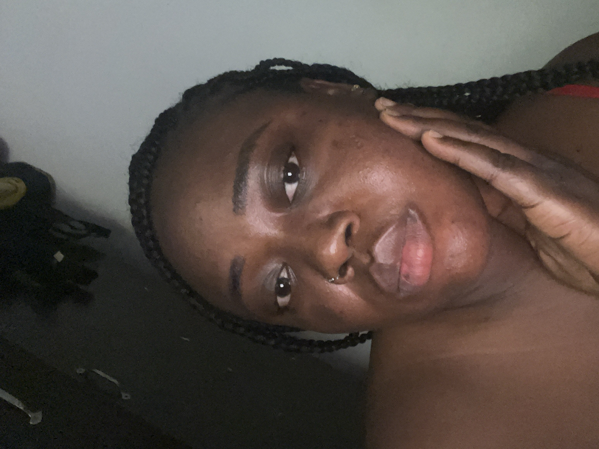 A close-up portrait features a person with a dark skin tone, rotated approximately 90 degrees clockwise, suggesting a spontaneous selfie. The individual has dark, intricately braided hair (box braids or similar style) framing their face and visible along the top and right side of the frame. Their dark eyes look directly at the viewer, conveying a neutral and engaging expression.
A small silver hoop nose ring is visible in their right nostril. Their lips are full and appear slightly parted, exhibiting a natural pinkish hue with some highlights. The skin shows natural texture and a subtle sheen. One hand is gently placed on the left side of their face, with fingers extending towards the temple. The nails are short and natural.
The setting appears to be indoors with soft, subdued lighting, highlighting features like the forehead, cheek, and lips, while casting much of the background into indistinct darkness. A lighter, plain surface is visible in the upper left quadrant of the image. The overall mood is intimate and contemplative. The location context provided is Abuja, Nigeria. No visible text is present in the image.