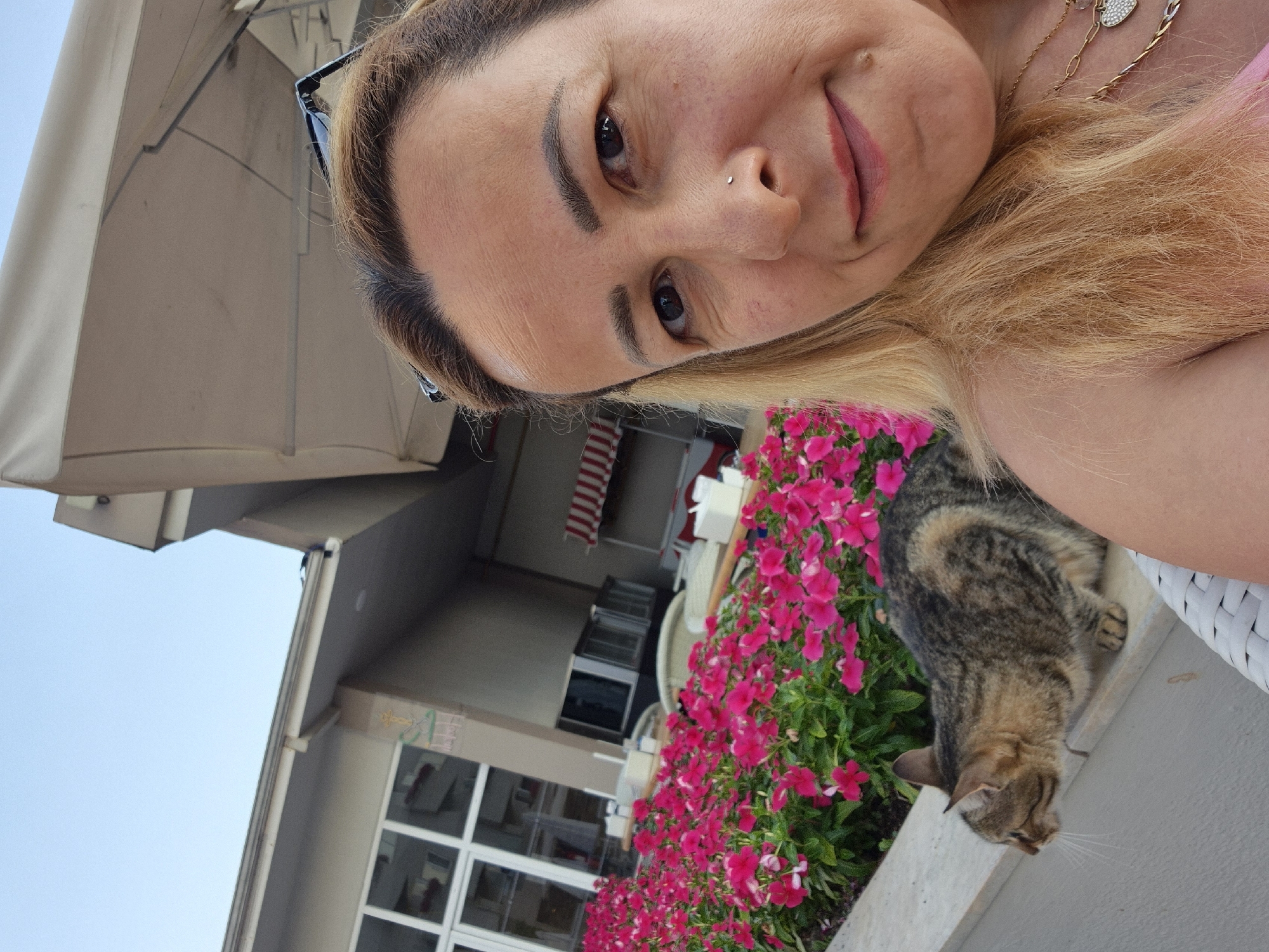 A woman with blonde hair and a nose ring is taking a selfie outdoors. She is wearing a pink top and a necklace. Next to her is a tabby cat, sitting on a ledge. Behind them is a building with large windows and a row of vibrant pink flowers. A large, light-colored awning is visible above the woman. The sky is clear and blue.