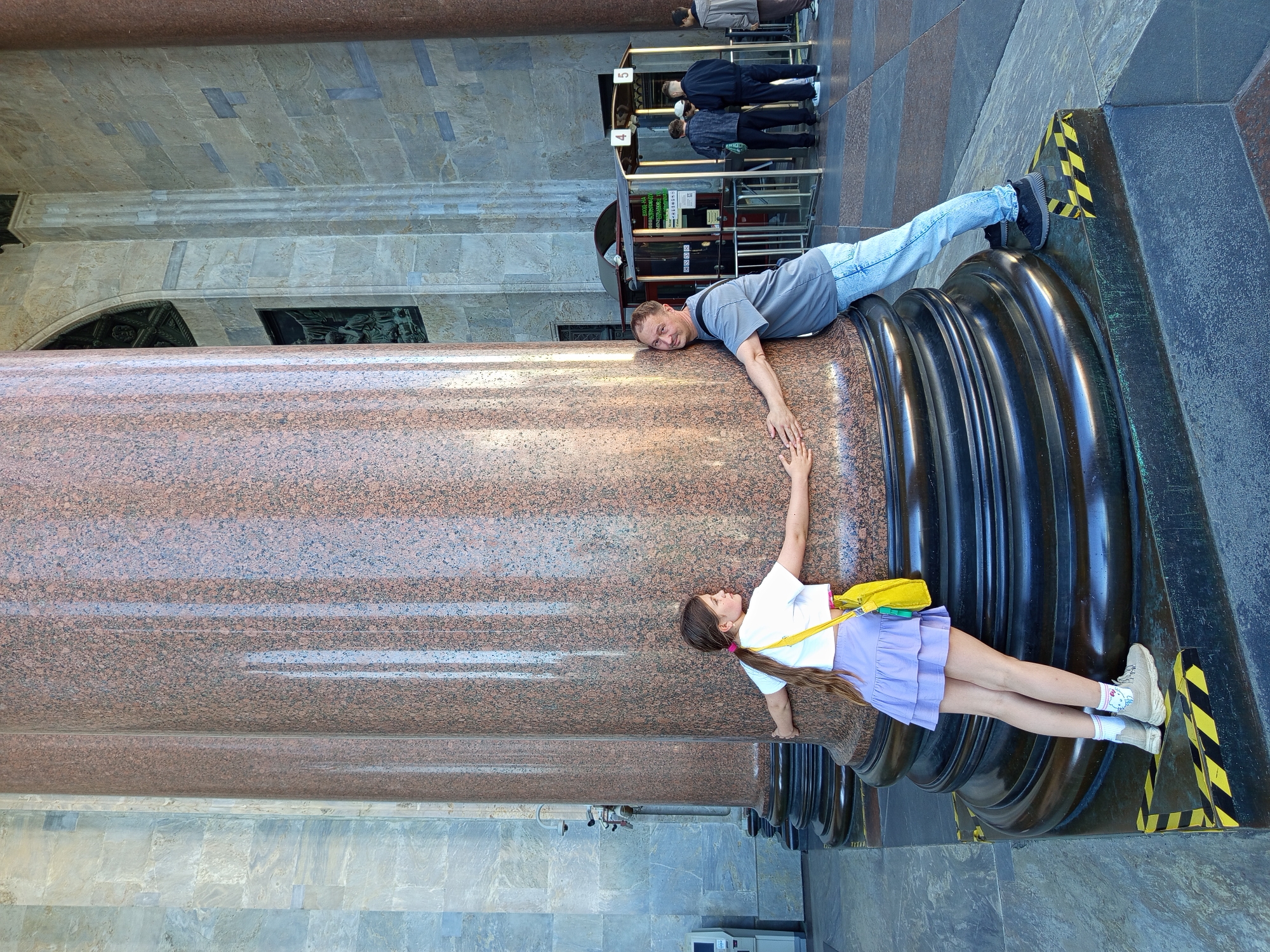 The image captures a father and daughter embracing a large, fluted granite column in front of the Kazan Cathedral in Saint Petersburg, Russia. The man, wearing a grey t-shirt and blue jeans, leans into the column with his arms wrapped around it, looking towards the camera with a smile. The daughter, with her long brown hair in a ponytail, wears a white t-shirt, a lavender ruffled skirt, and a yellow crossbody bag. She also has her arms around the column, facing away from the camera. The column is made of reddish-brown granite with a polished surface, and it stands on a dark, tiered base. In the background, people can be seen near ticket booths and entrances to the cathedral. The architecture suggests a grand, classical style, typical of Saint Petersburg's prominent landmarks. The lighting indicates a bright, sunny day.
