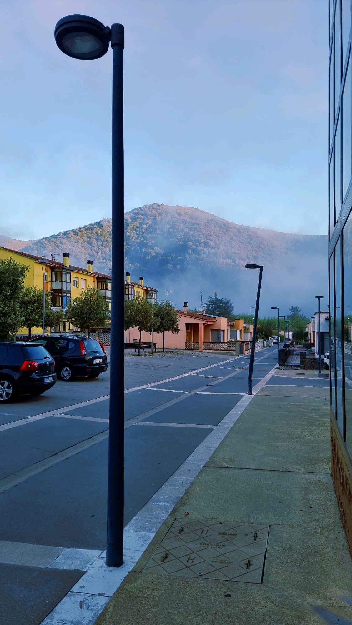 The image depicts a street scene in Sant Joan Les Fonts, Spain, captured at an angle from the sidewalk, looking down a street towards a distant misty mountain. In the foreground, a dark lamppost stands tall, to the right of which is a modern building with reflective glass windows. The street itself is paved with asphalt, marked with white lines forming parking spaces. Several cars are parked along the left side of the street, including two black cars in the foreground. Across the street, a row of low-rise buildings, predominantly yellow and peach-colored, are visible, with trees lining the sidewalk in front of them. A gentle mist hovers over the lower slopes of the tree-covered mountain in the background, under a pale blue sky. The overall atmosphere is calm and quiet, suggesting an early morning or late evening setting.