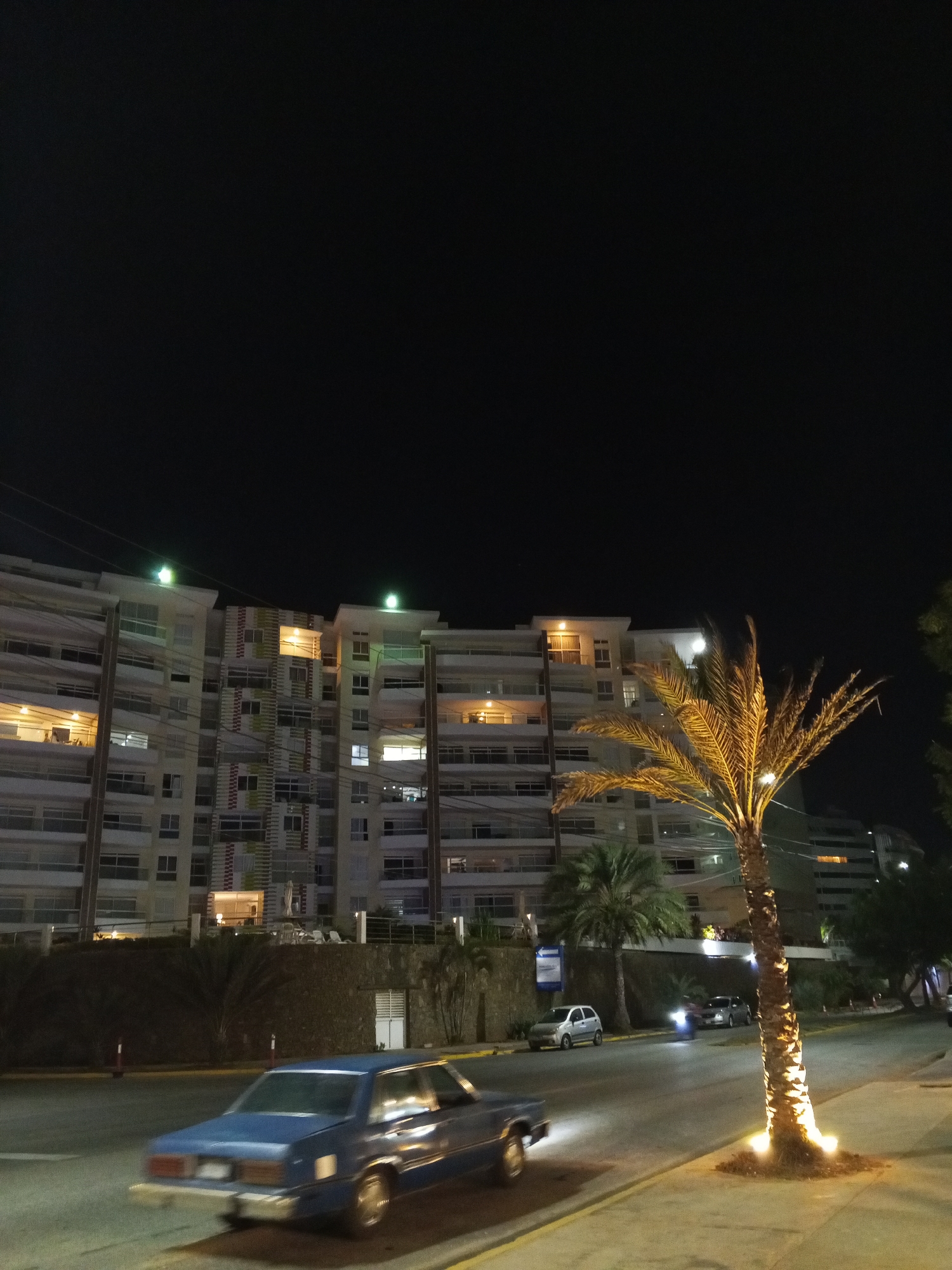 The image shows a nighttime urban scene in Pampatar, Venezuela. A multi-story residential building with lighted windows is in the background. A palm tree, illuminated from below, stands on the right sidewalk. A blue car is moving on the street, with motion blur. Other parked cars and vegetation are visible along the road. The sky is dark, suggesting it is night.