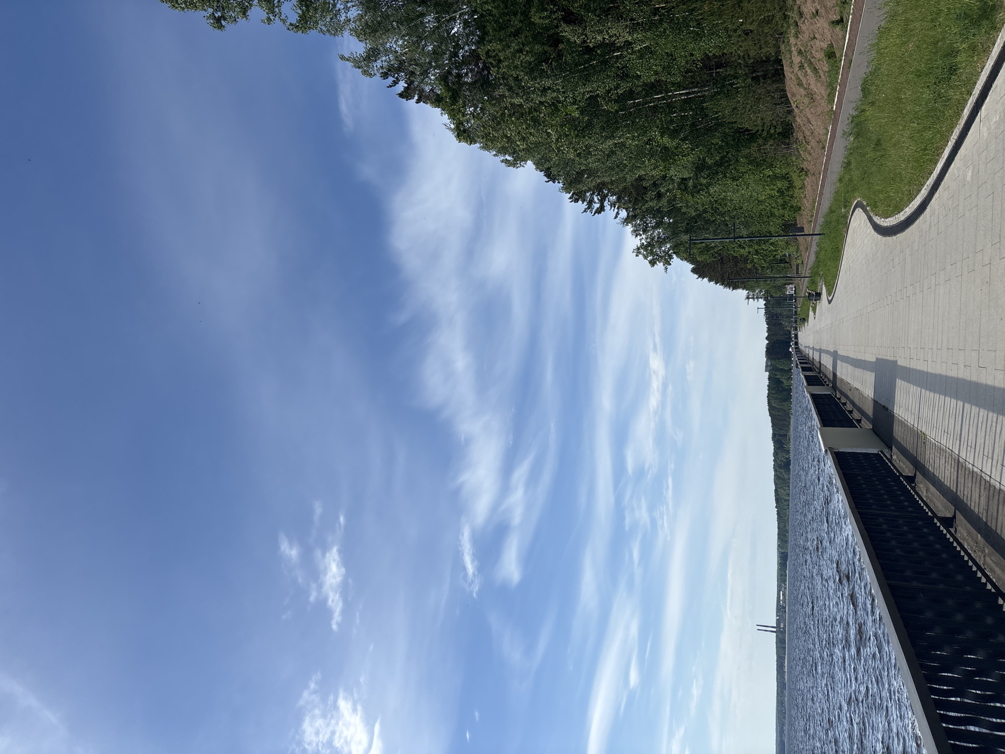 The image depicts a waterfront scene in Chaikovsky, Russia. The view is of the embankment, adjacent to the Kama River. The sky is blue with streaks of clouds. A paved walkway runs along the top right of the frame, bordered by a grassy area and trees. A metal railing separates the walkway from the water. The river is calm with a dark blue hue. A structure is visible in the distance on the river. Black lampposts are positioned along the walkway.