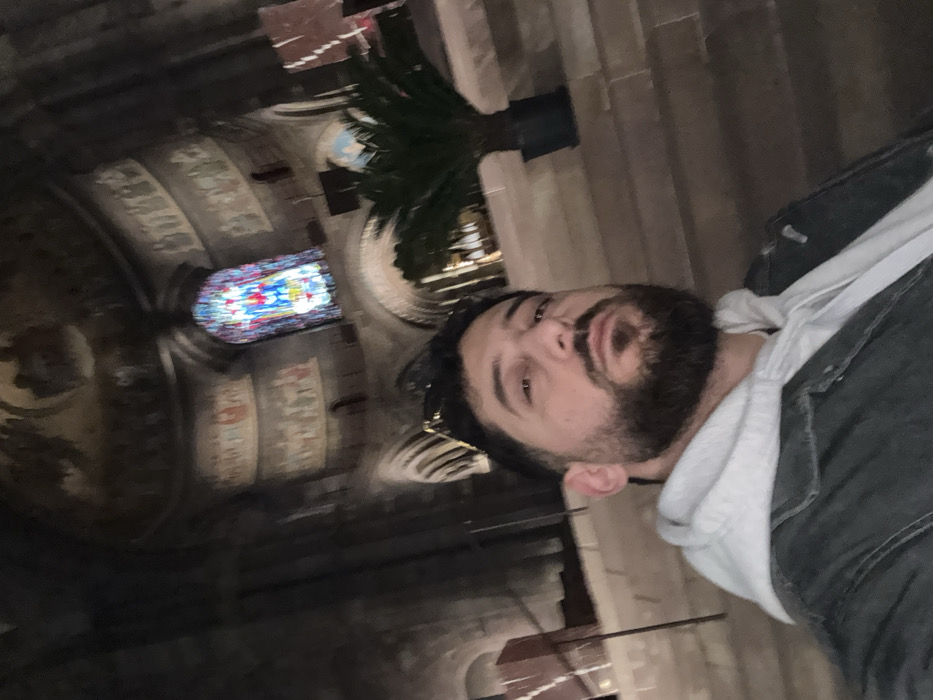An adult male is captured in a selfie-style photograph within the Cathédrale Notre-Dame de Strasbourg, France. The subject, in the foreground, has dark hair, a full beard, and is looking directly towards the camera, with sunglasses resting on top of his head. He is wearing a light grey hooded sweatshirt beneath a dark, possibly denim, jacket.
The background reveals the dimly lit interior of the cathedral, characterized by imposing dark stone walls and a high, vaulted ceiling with rounded arches, indicative of Romanesque architecture. Upper wall sections and the ceiling are adorned with multi-figure murals or frescoes depicting various scenes in muted tones. A brightly illuminated rectangular stained-glass window, primarily displaying blue, red, and yellow hues within a detailed depiction of a figure or scene, is prominent within a deep archway in the background. To the right, a large potted plant with green fronds is visible on a raised stone platform. The environment suggests a grand, historic religious structure with solemn lighting.