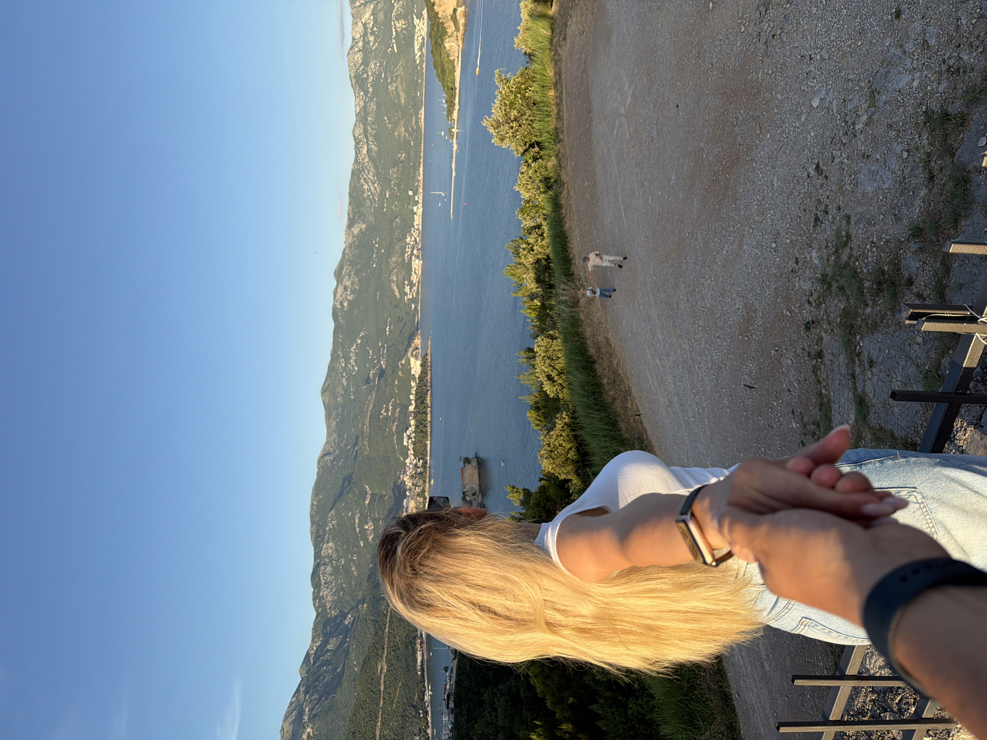 An over-the-shoulder, POV shot captures a woman with long blonde hair, facing away from the camera and holding the hand of the unseen photographer. She is wearing a white ribbed tank top and light blue denim jeans. She has a smartwatch on her left wrist. The scene is set outdoors with a scenic view of the Adriatic Sea and a coastline dotted with buildings, backed by a range of mountains under a clear blue sky. In the mid-ground, two figures are walking away from the camera on a gravel path. The foreground features a black metal railing. This vista is consistent with the area around Budva, Montenegro, possibly from a vantage point overlooking the town and its bay. The lighting suggests it is late afternoon or early evening, with the sun casting a warm glow.
