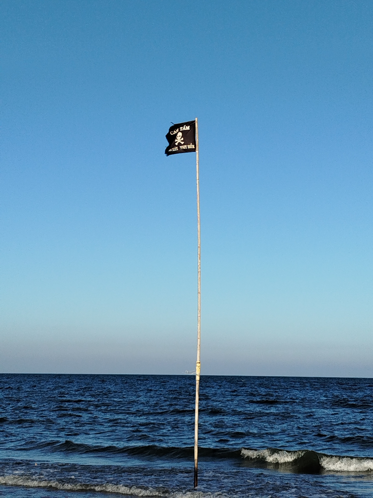 A black pirate flag with a skull and text flies on a tall pole near the ocean. The scene is sunlit with a clear blue sky above the ocean. The ocean water is dark with small waves visible near the shore. This is most likely a beach in the city of Ho Chi Minh City, Vietnam.